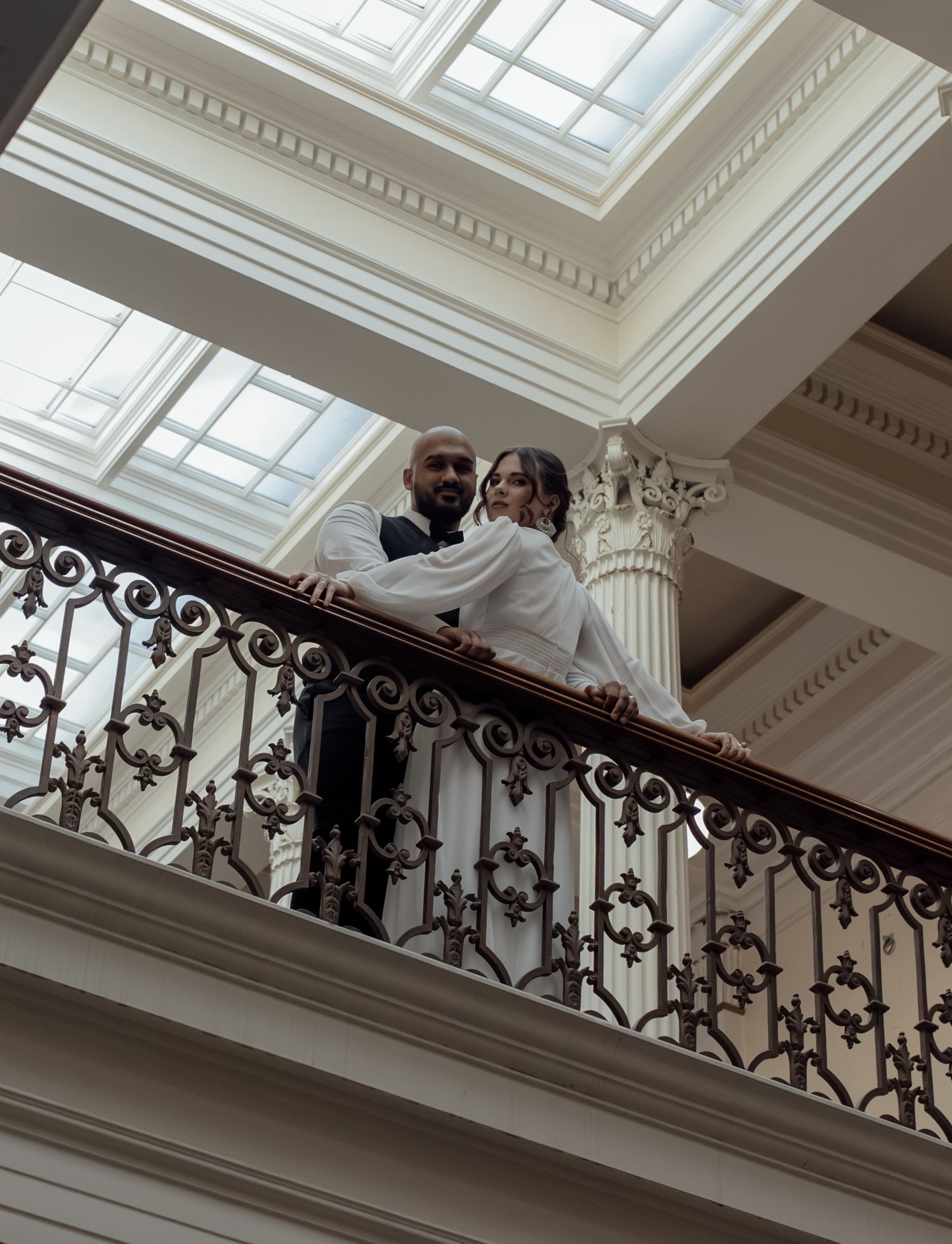 A couple poses on an elegant staircase with ornate iron railings and white columns under a skylit ceiling.