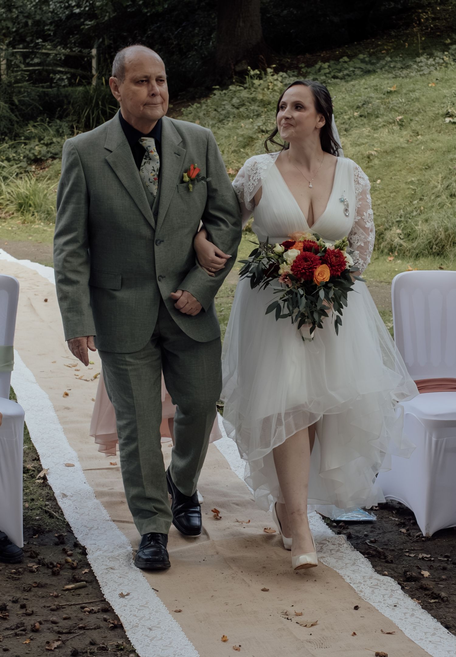 A wedding couple walks down an outdoor aisle together, with the bride holding a vibrant red and burgundy bouquet.