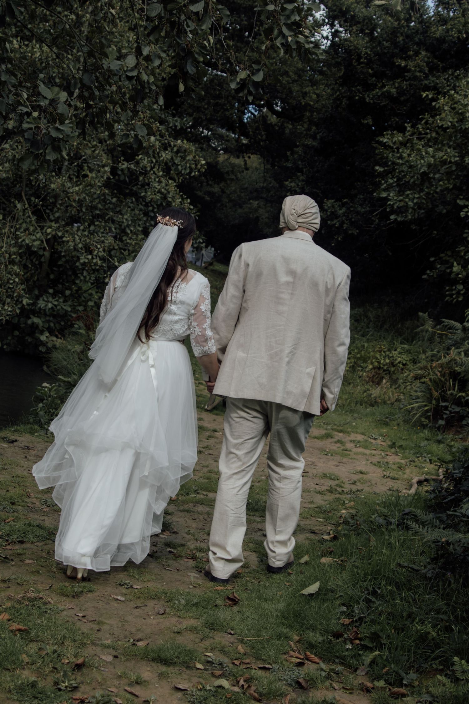 A couple in wedding attire walks along a wooded path, with the bride's white dress flowing behind them.
