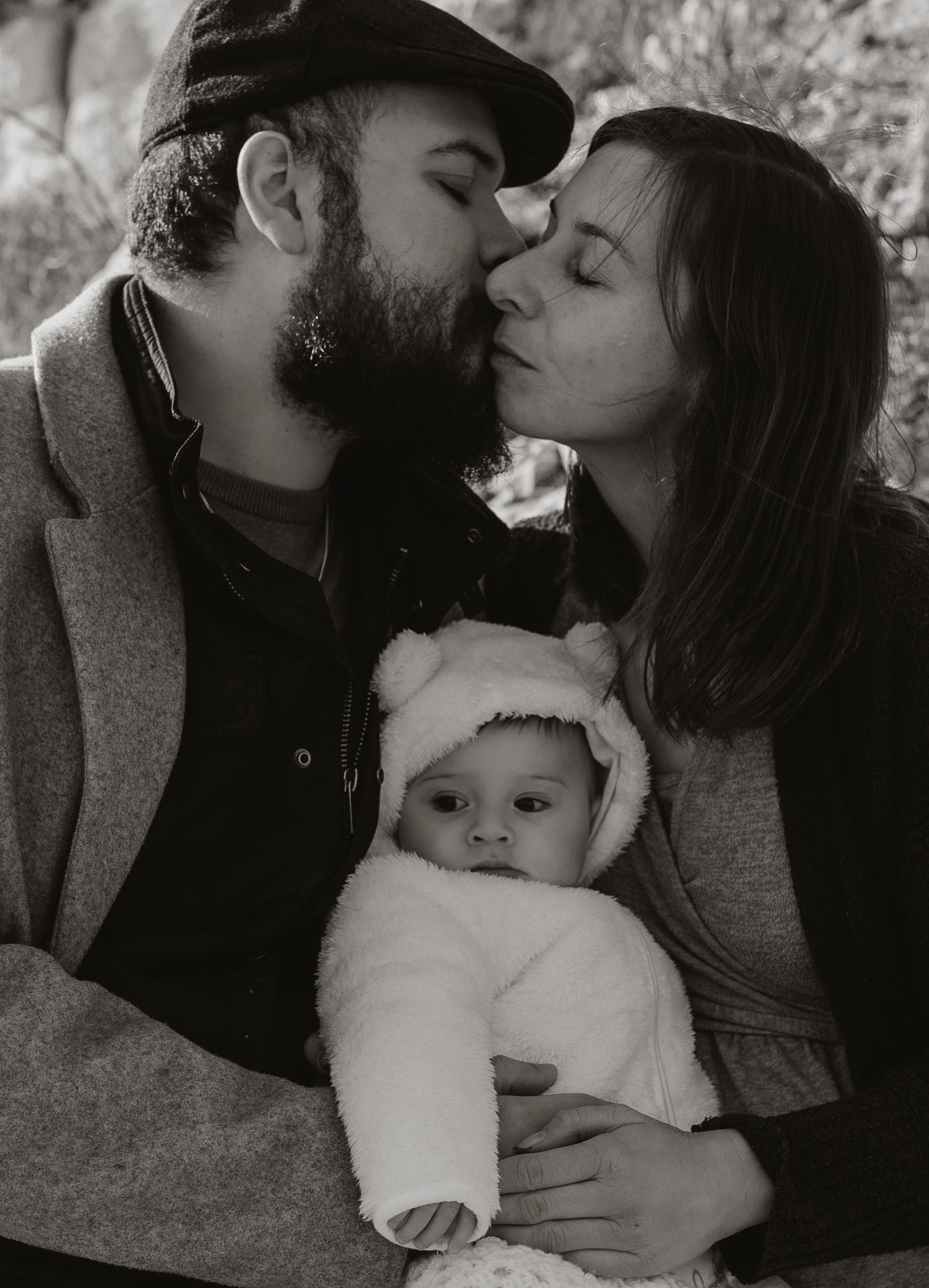 Black and white portrait of a couple sharing an intimate moment with their baby bundled in white.