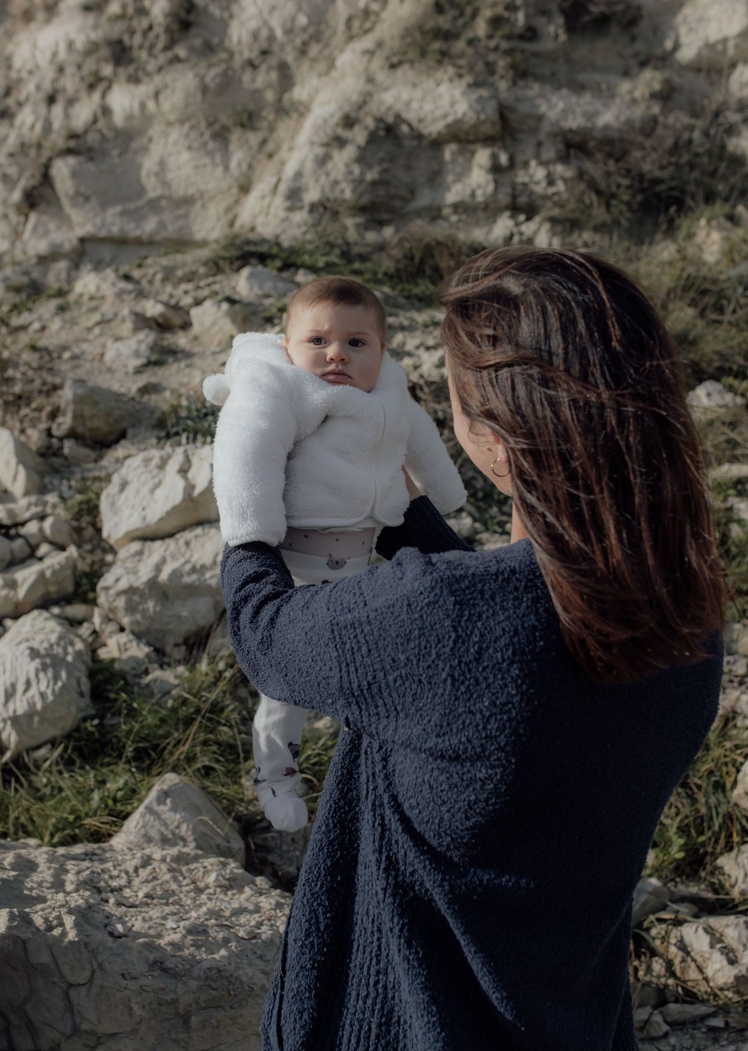 A parent in a dark sweater holds and comforts a small baby against a stone wall backdrop.