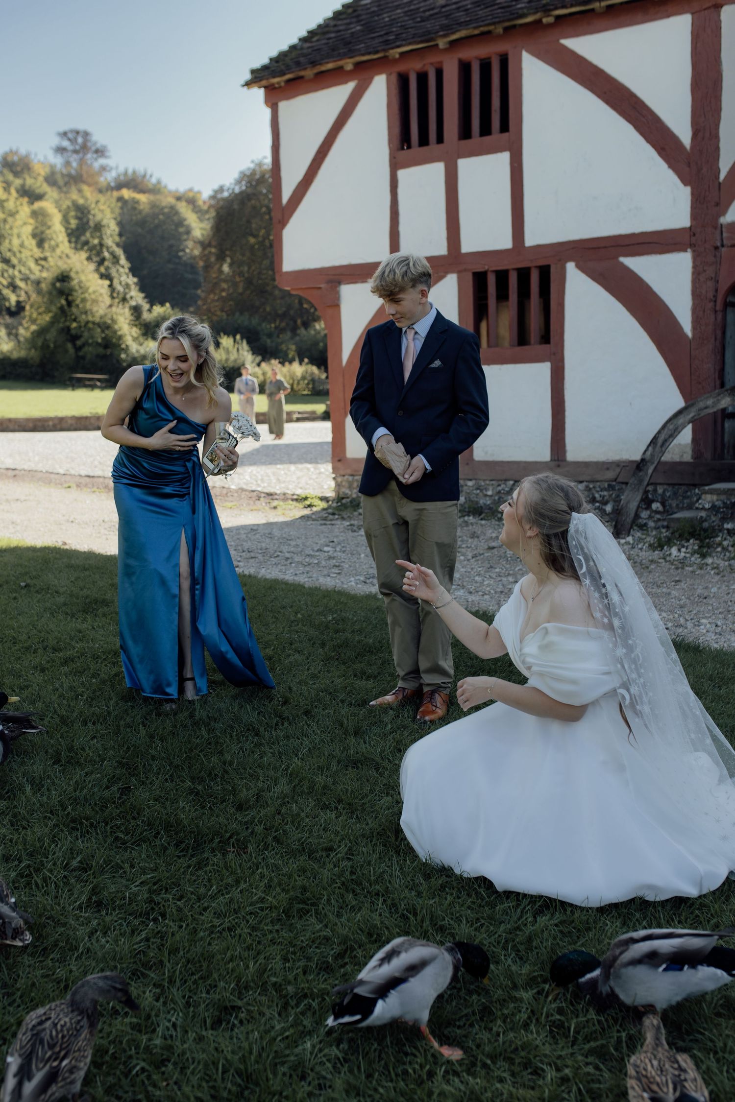 A couple in wedding attire shares a playful moment outdoors near a rustic white building with timber framing.
