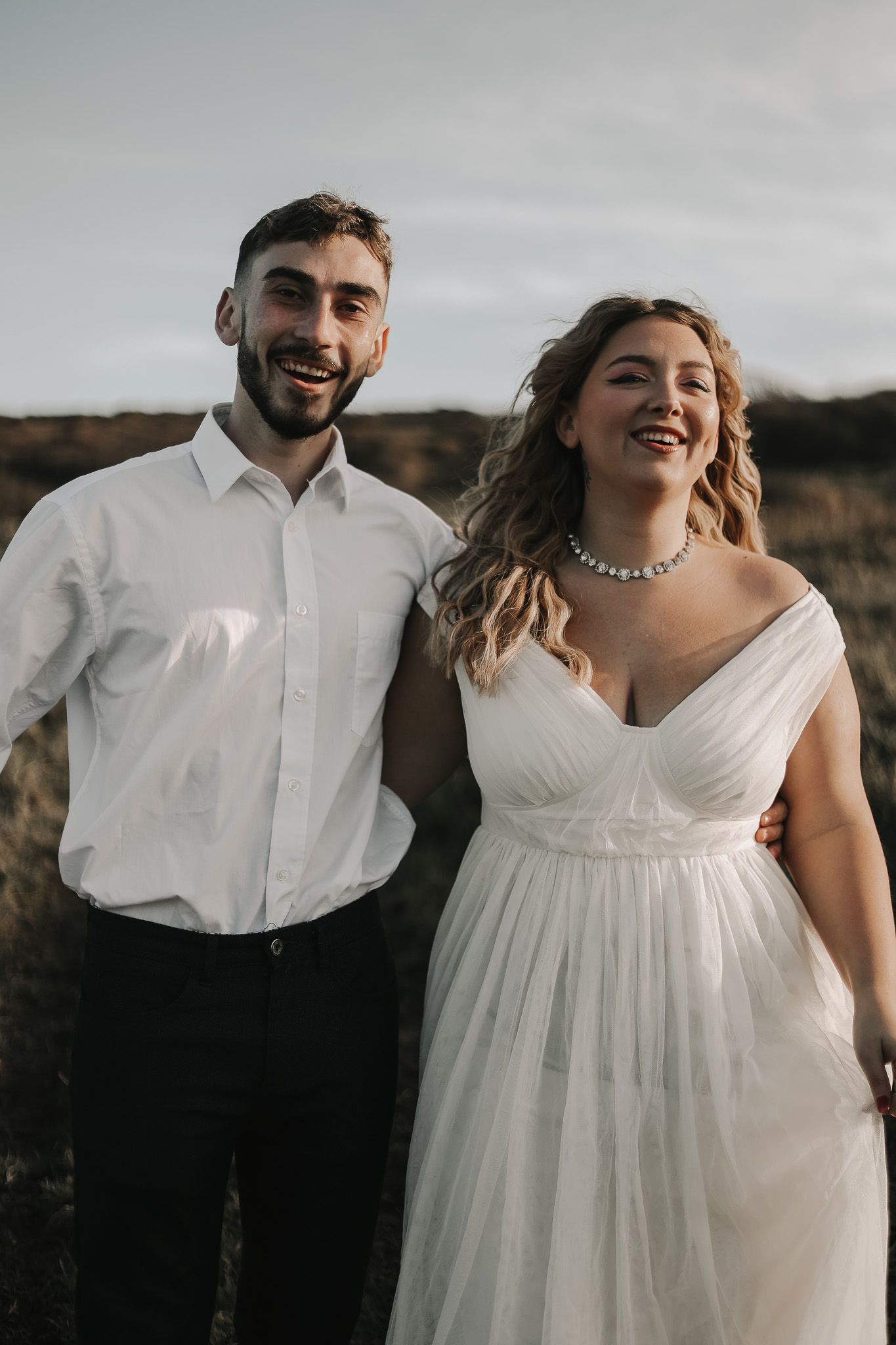 A couple poses together outdoors for wedding photos in a white dress and dress shirt against a moody sky.