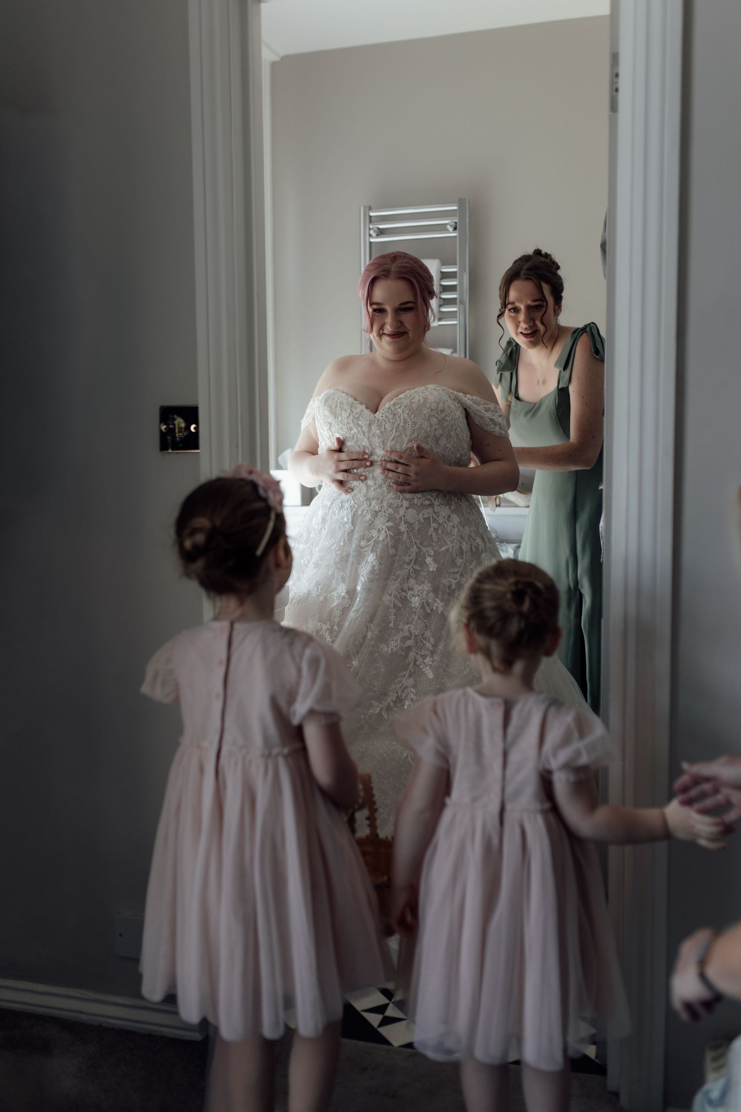 A bride in a white wedding dress stands with two young flower girls in pink dresses in a bridal preparation room.