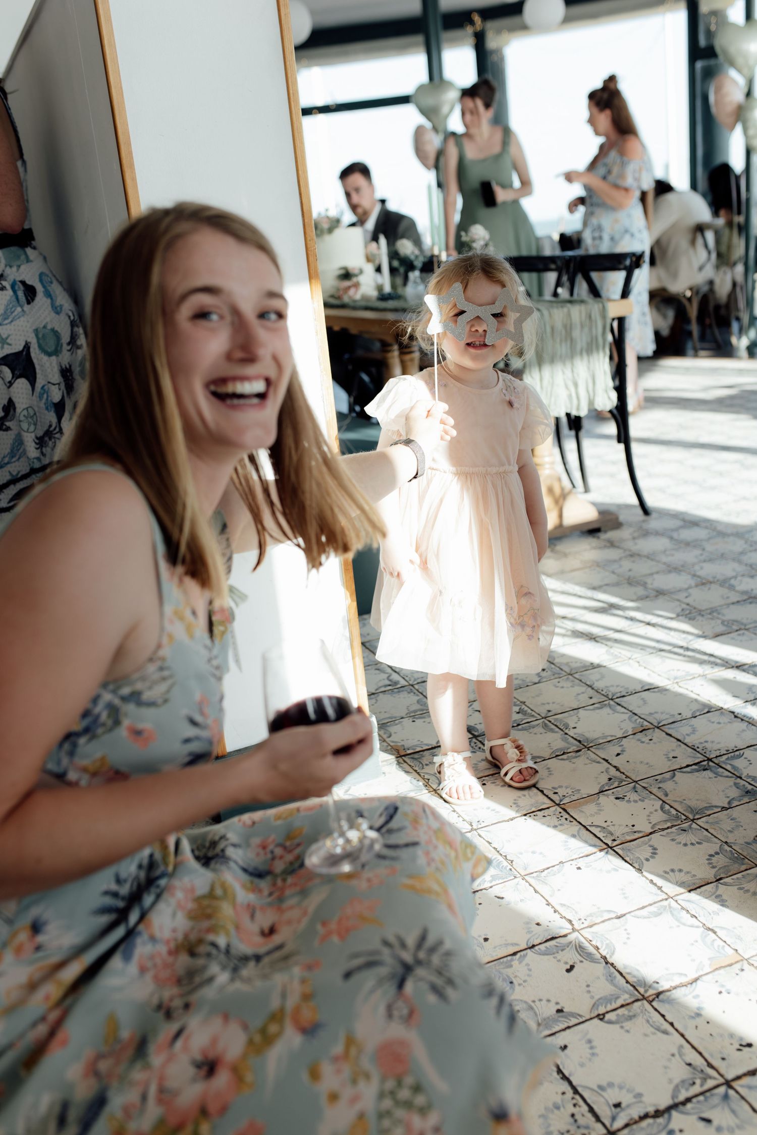 A festive indoor celebration with people in floral dresses and a small child in a light pink dress on a tiled floor.