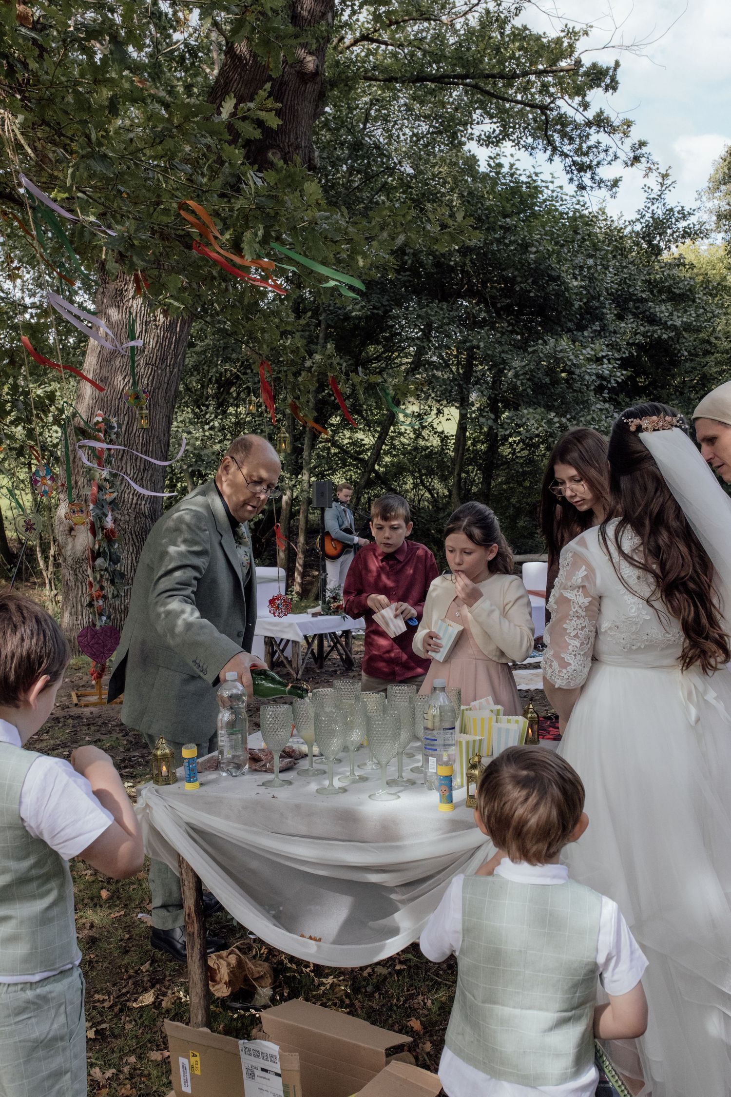People gather around a table at an outdoor event with drinks and decorations under tree branches.