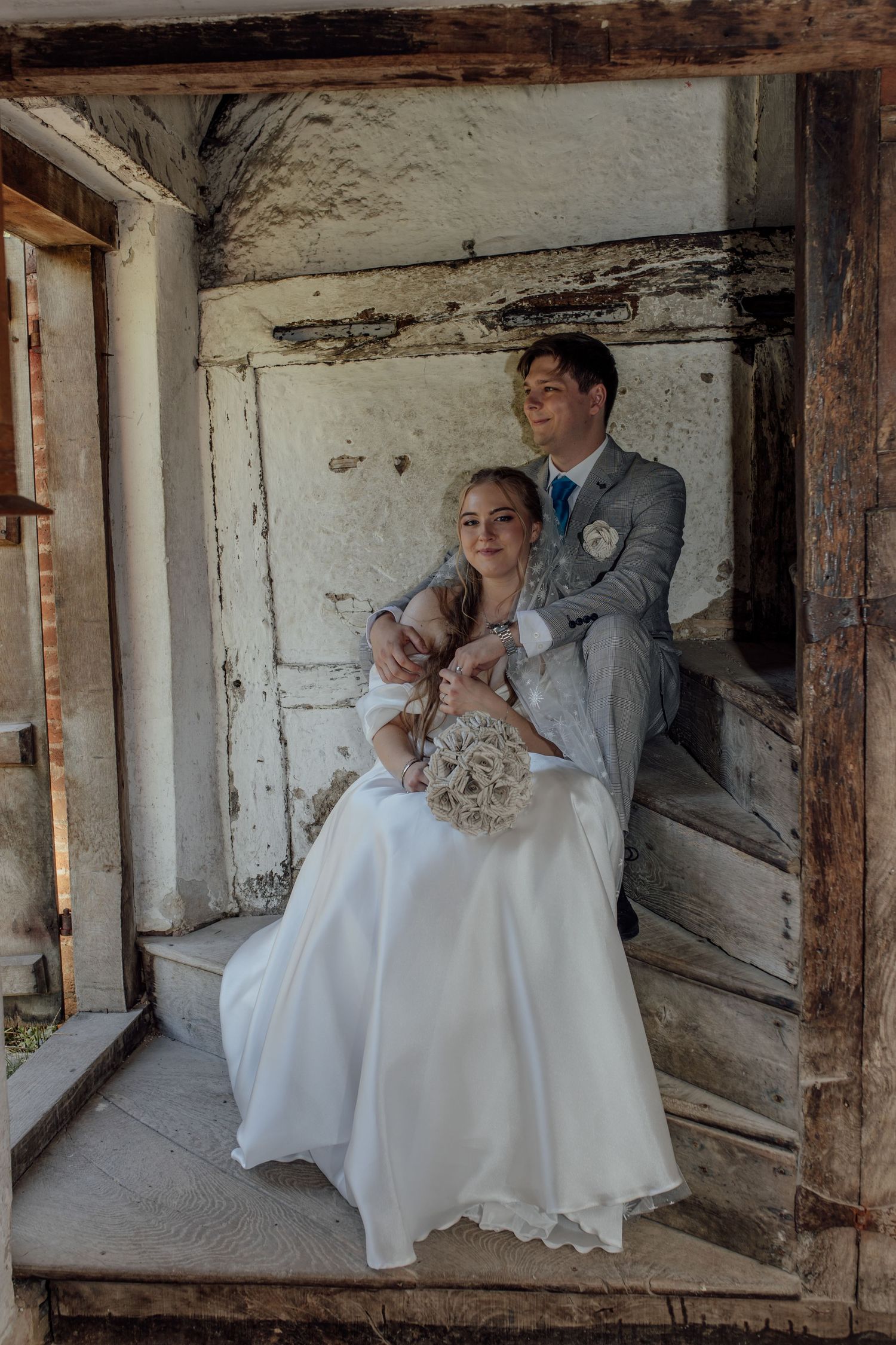 Romantic wedding couple poses together in rustic barn setting with exposed wooden beams and white stone walls.