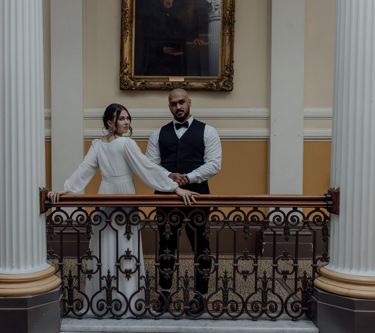 A couple shares a romantic moment on a balcony overlooking an elegant white-columned interior space.