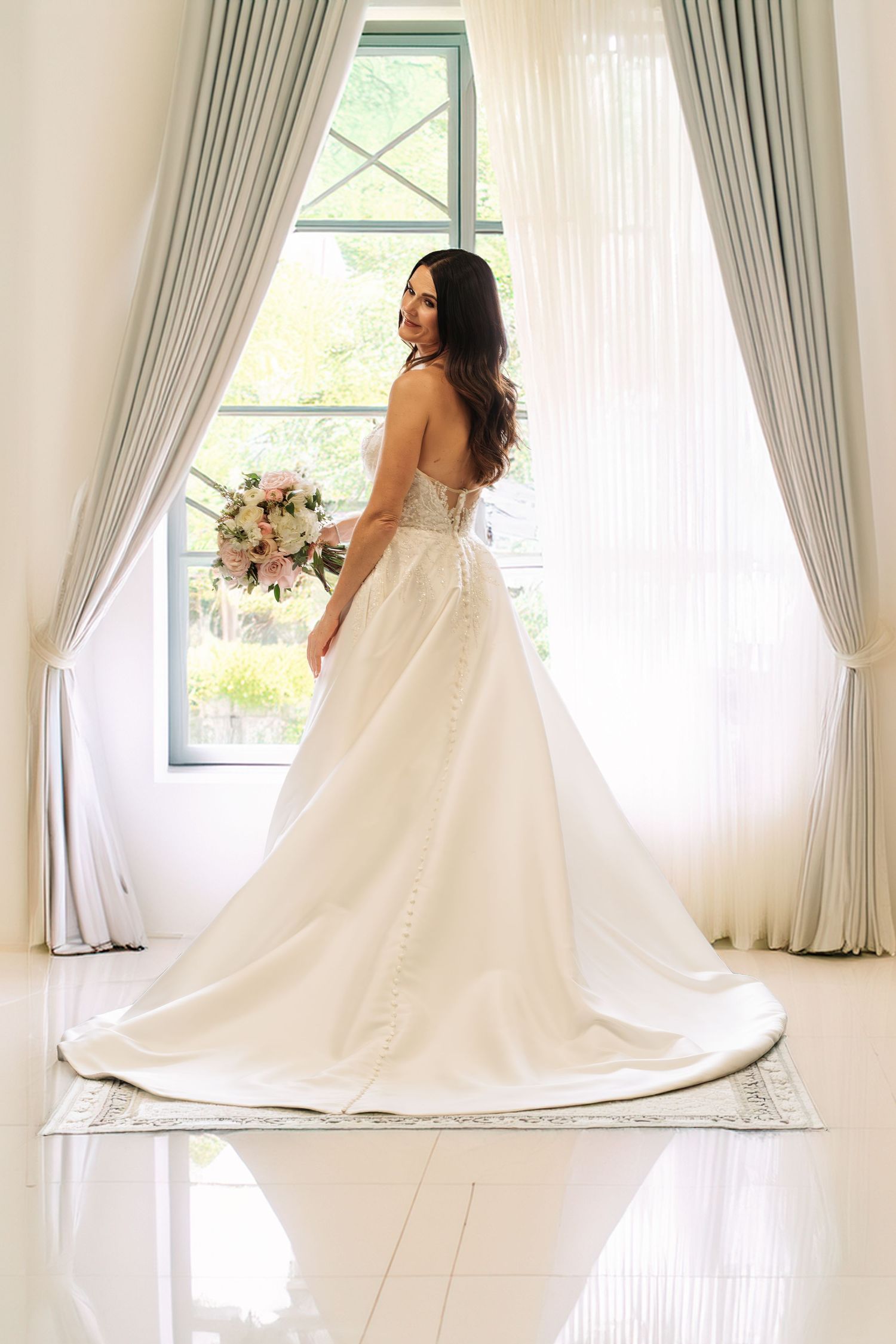 “Bride looking back and smiling while holding her bouquet, framed by large bright windows and flowing white curtains.”
