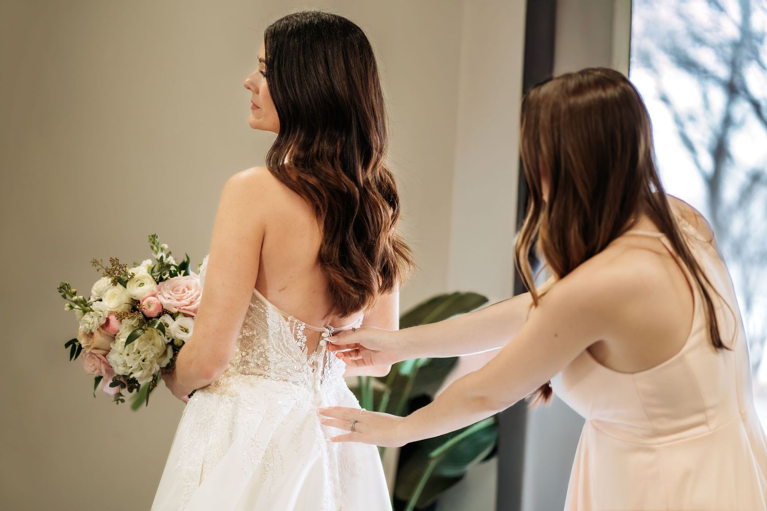 “Bridesmaid fixing the back of the bride’s gown during a quiet getting‑ready moment.”