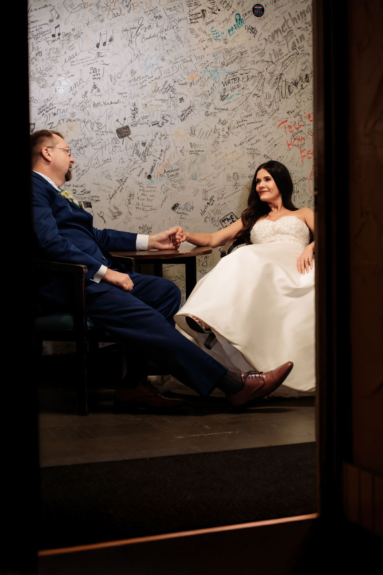 “Candid peek‑through doorway shot of a wedding couple sitting together and holding hands with a table between them.”