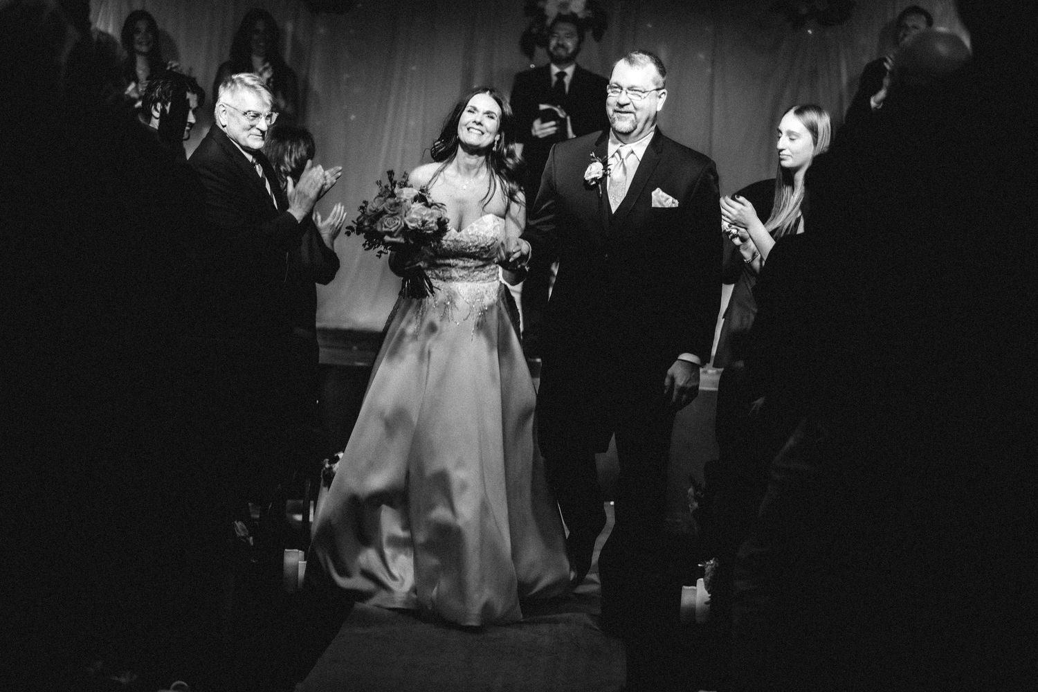 “Black and white photo of a newly married couple walking down the aisle and greeting guests during their recessional.”