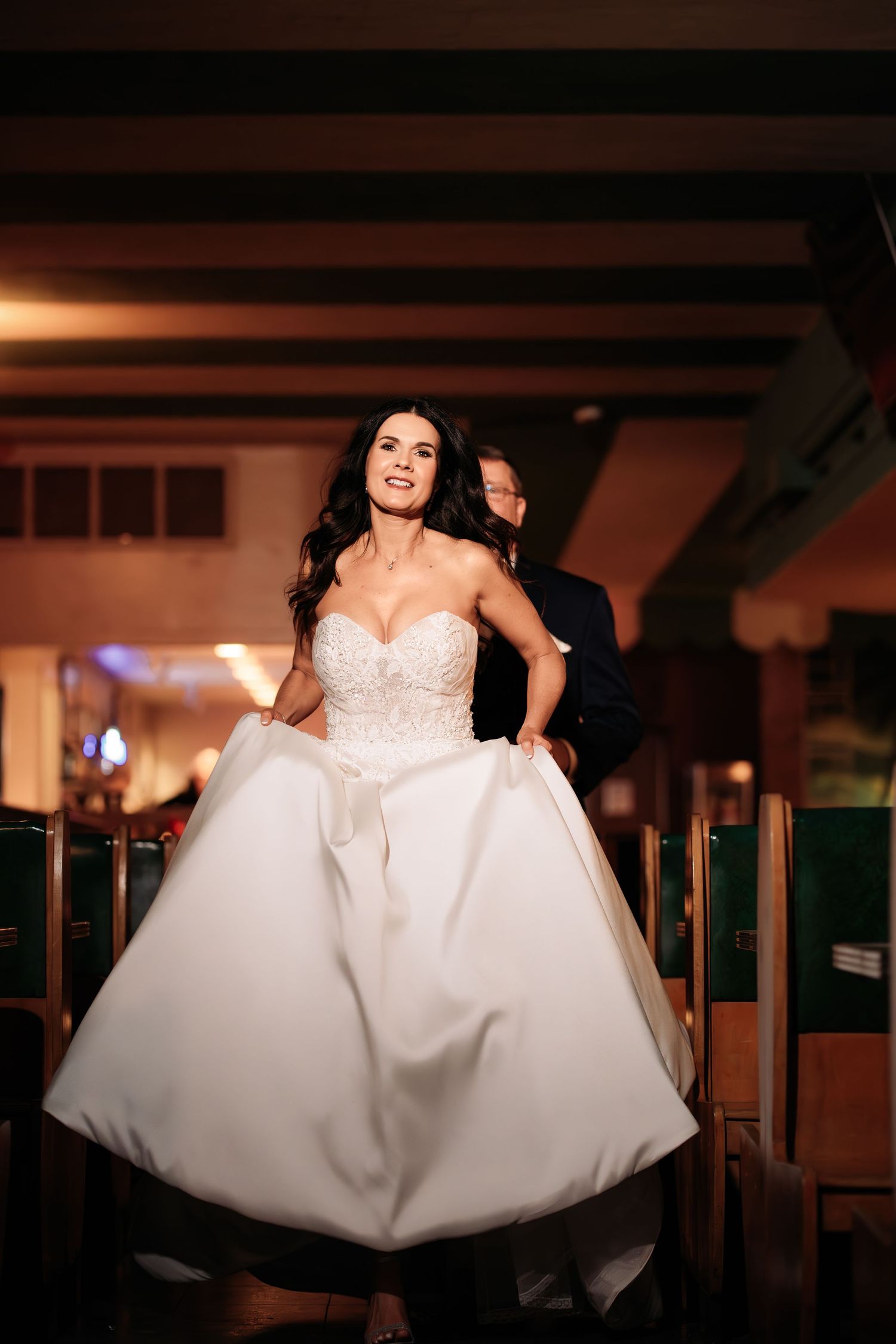 “Bride running in the foreground with the groom playfully chasing her in the aisle between banquet tables, moody candid moment.”