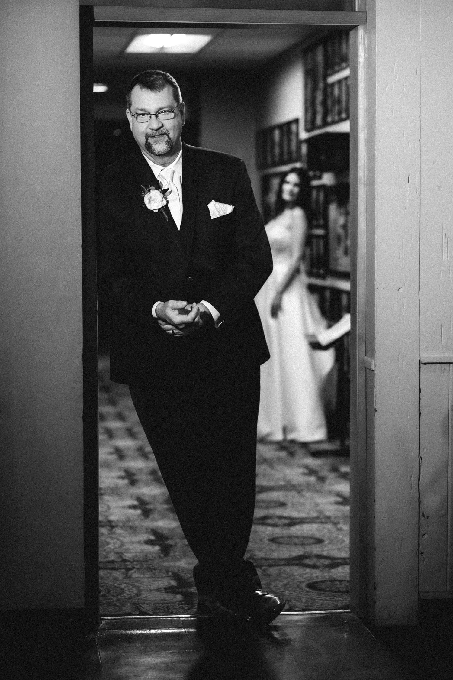 “Black and white portrait of a groom leaning on a door frame and looking at the camera, with the bride softly blurred in the background.”