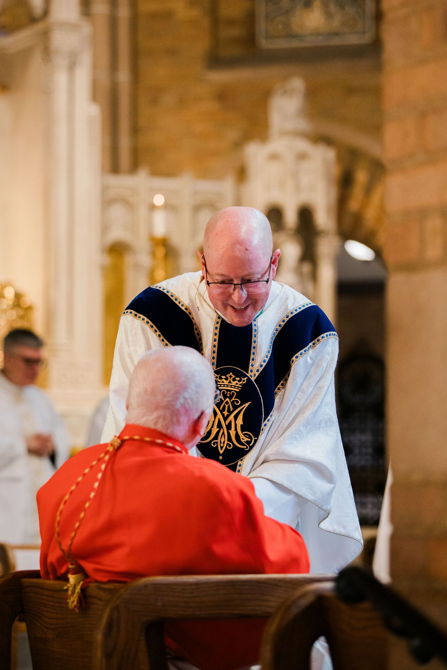A smiling Catholic priest in ornate vestments blesses a seated cardinal during a church ceremony.