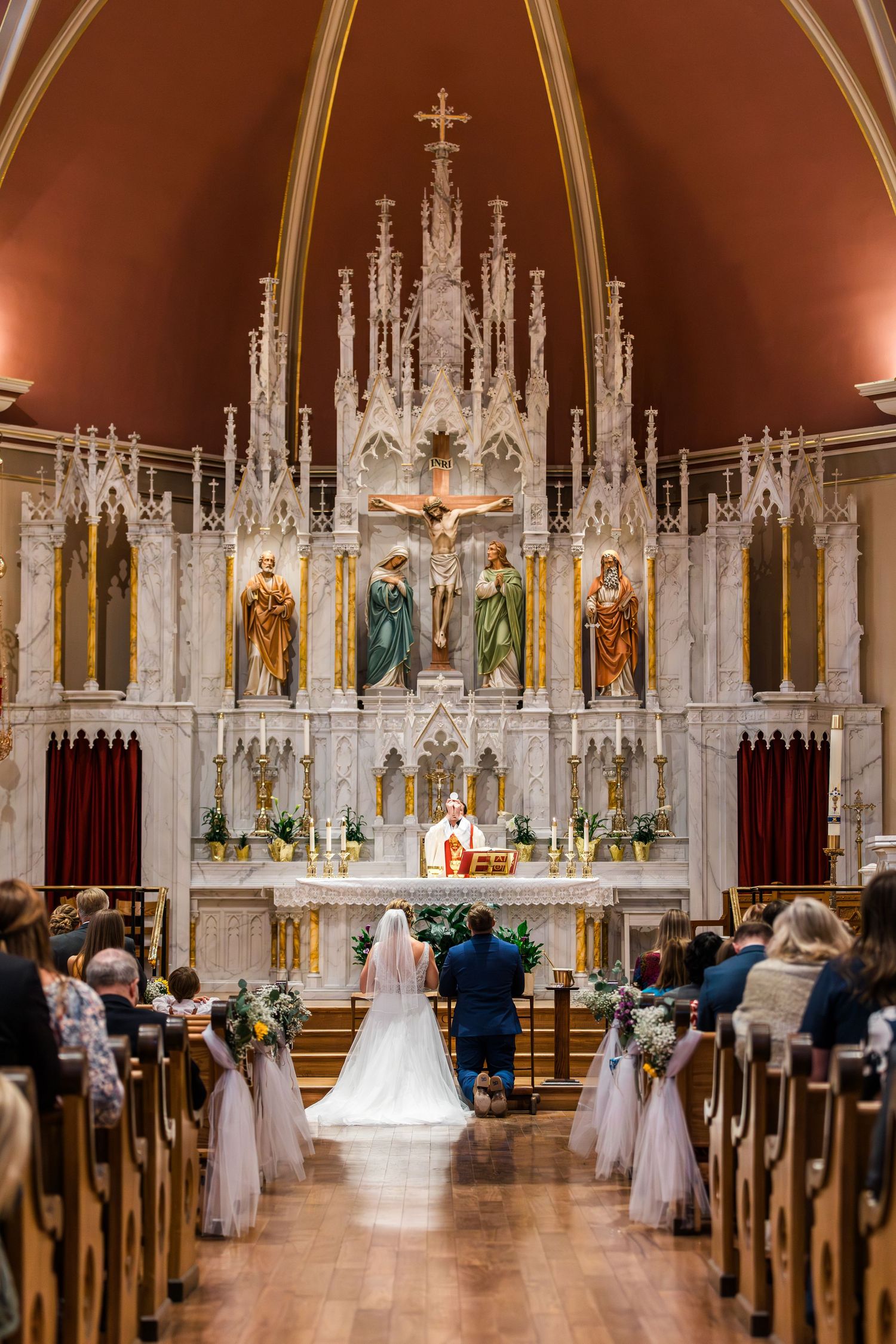 Bride and groom kneeling at ornate white altar during Catholic wedding ceremony in historic church.