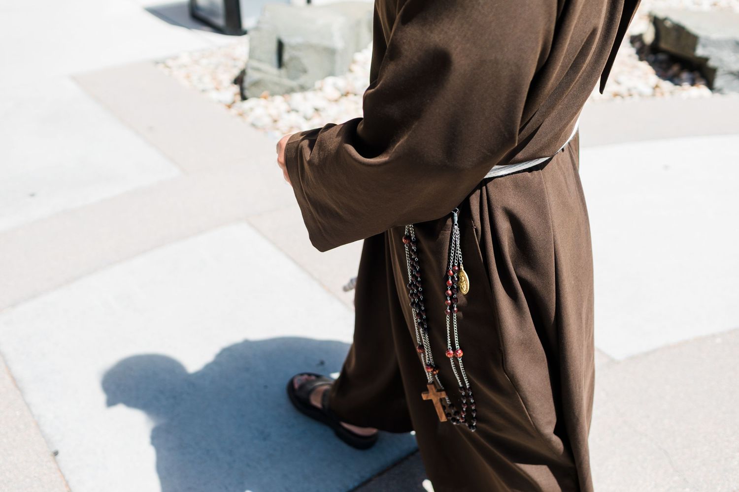 A Franciscan monk in brown habit walks outdoors, rosary beads hanging from his side, casting a shadow on the pavement.