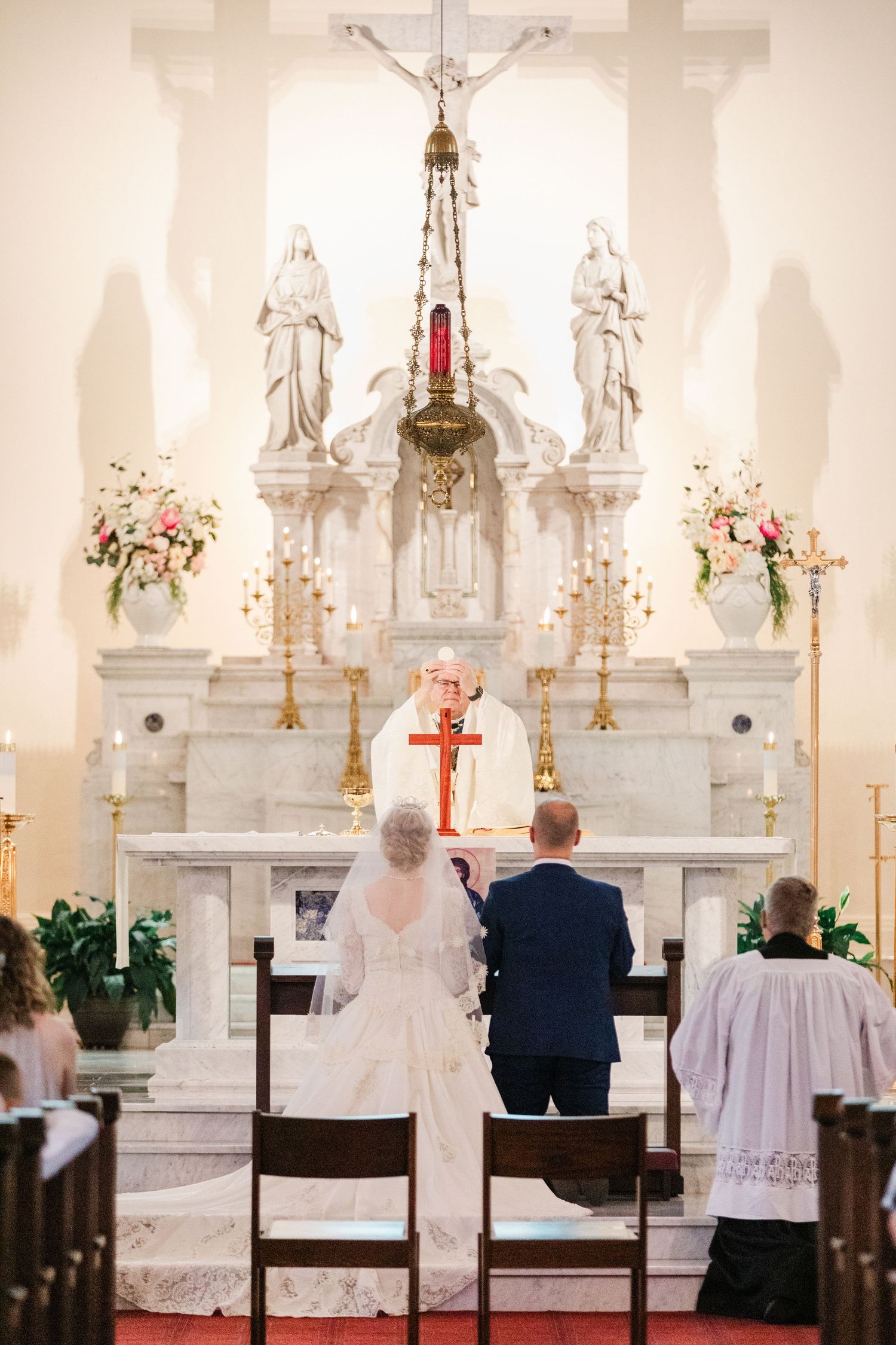 Bride in lace gown and groom kneel before priest at elegant white marble altar adorned with floral arrangements and candles.
