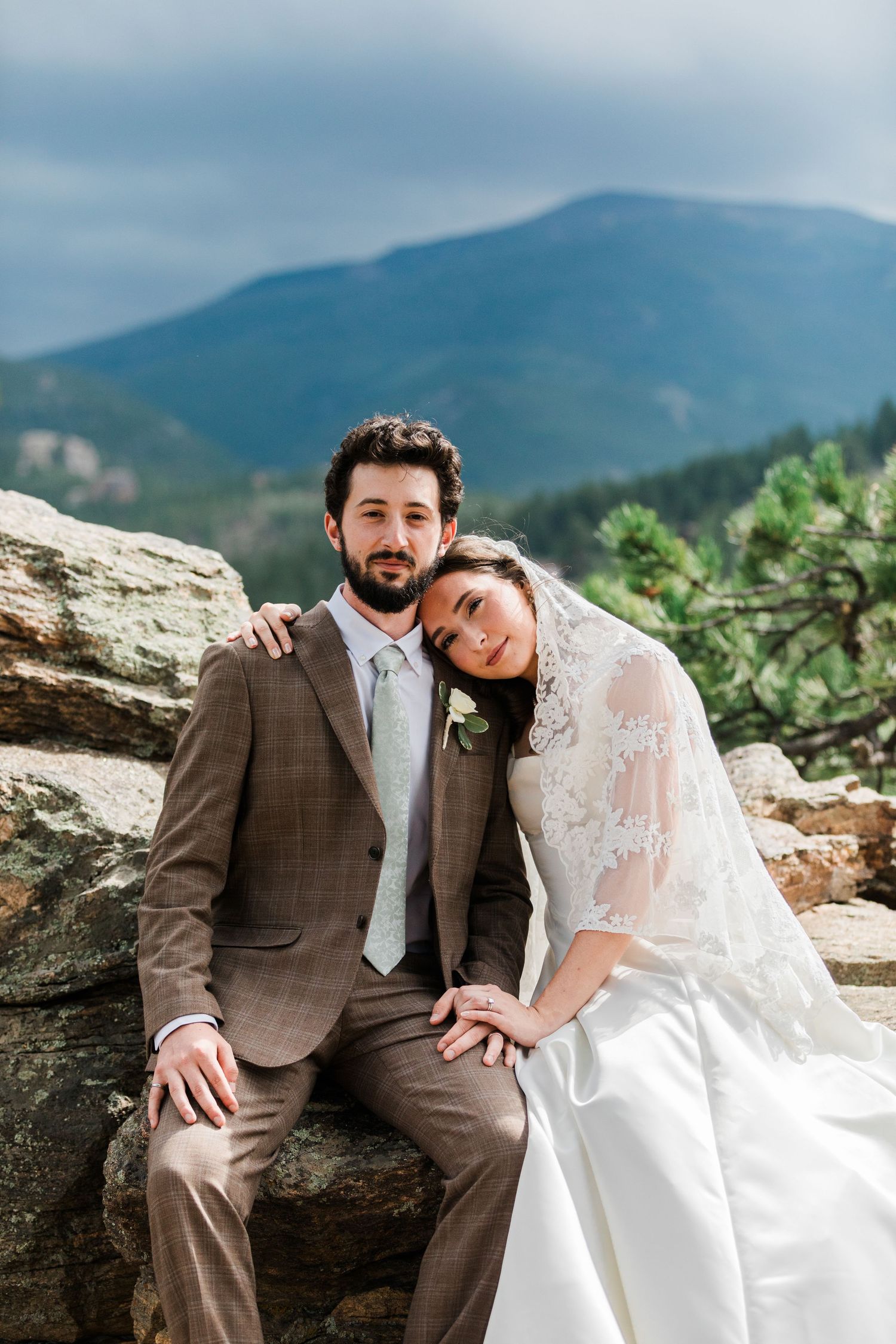 Bride and groom sitting on rocky ledge with mountain backdrop, bride in lace veil leaning on groom in brown suit.