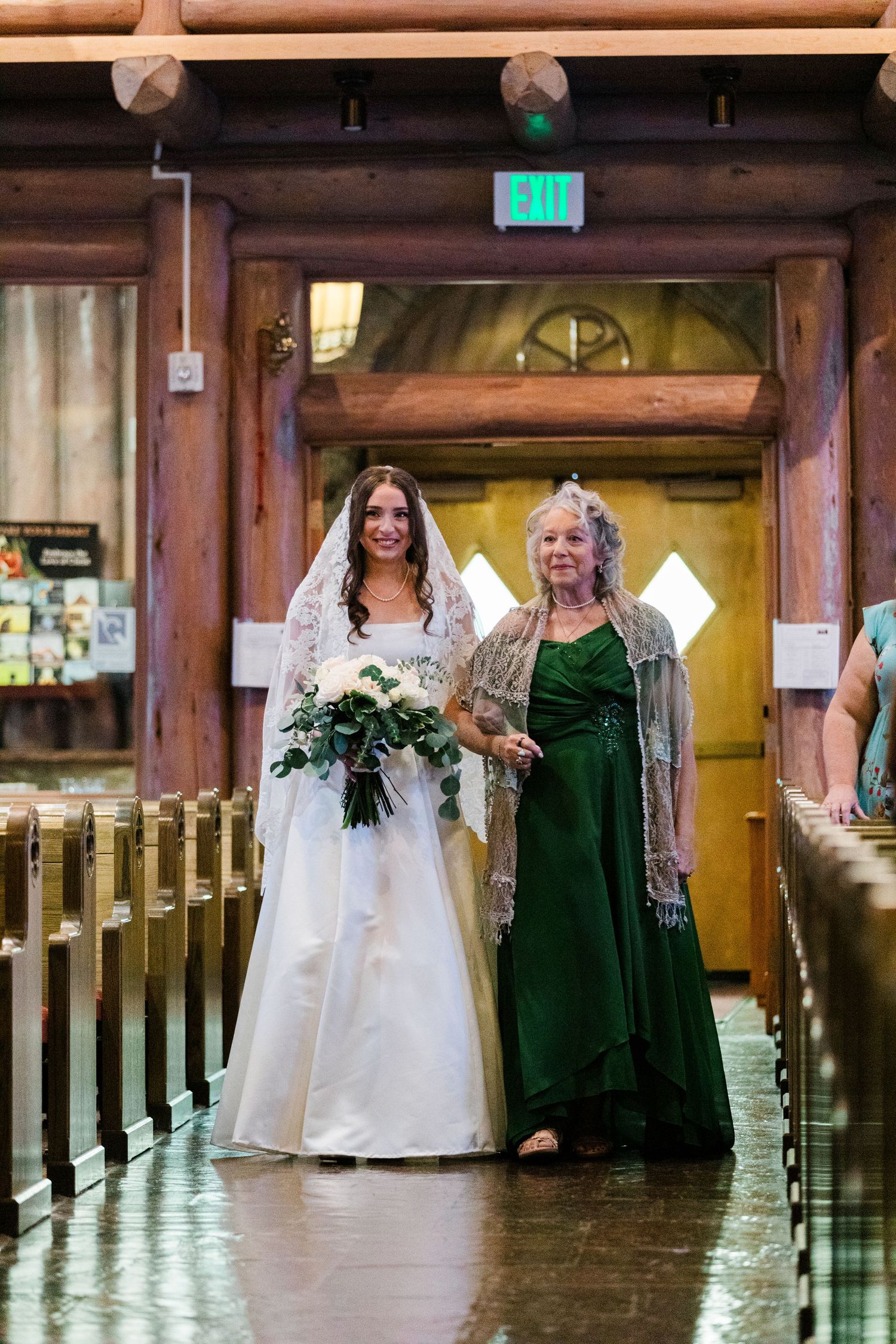 Bride in white gown and veil walks down church aisle with older woman in green dress during wedding ceremony.