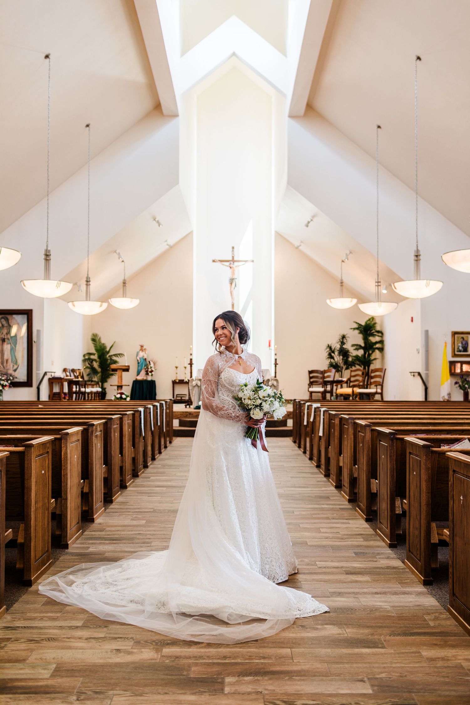 Bride in lace wedding gown stands in church aisle holding bouquet, with wooden pews and cross altar in background.