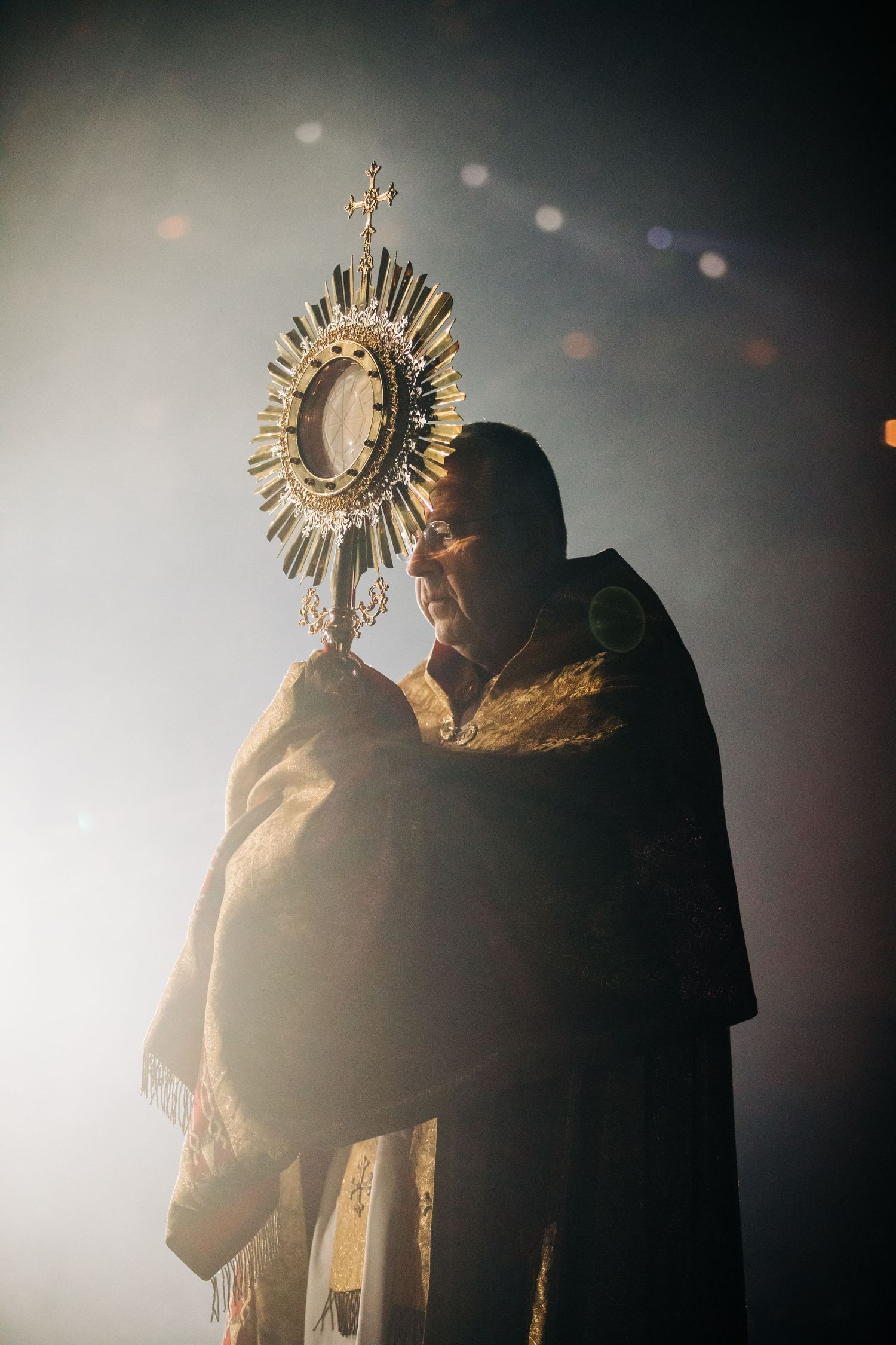 A dramatic silhouette of a religious figure holding a golden monstrance in atmospheric lighting during a Catholic ceremony.