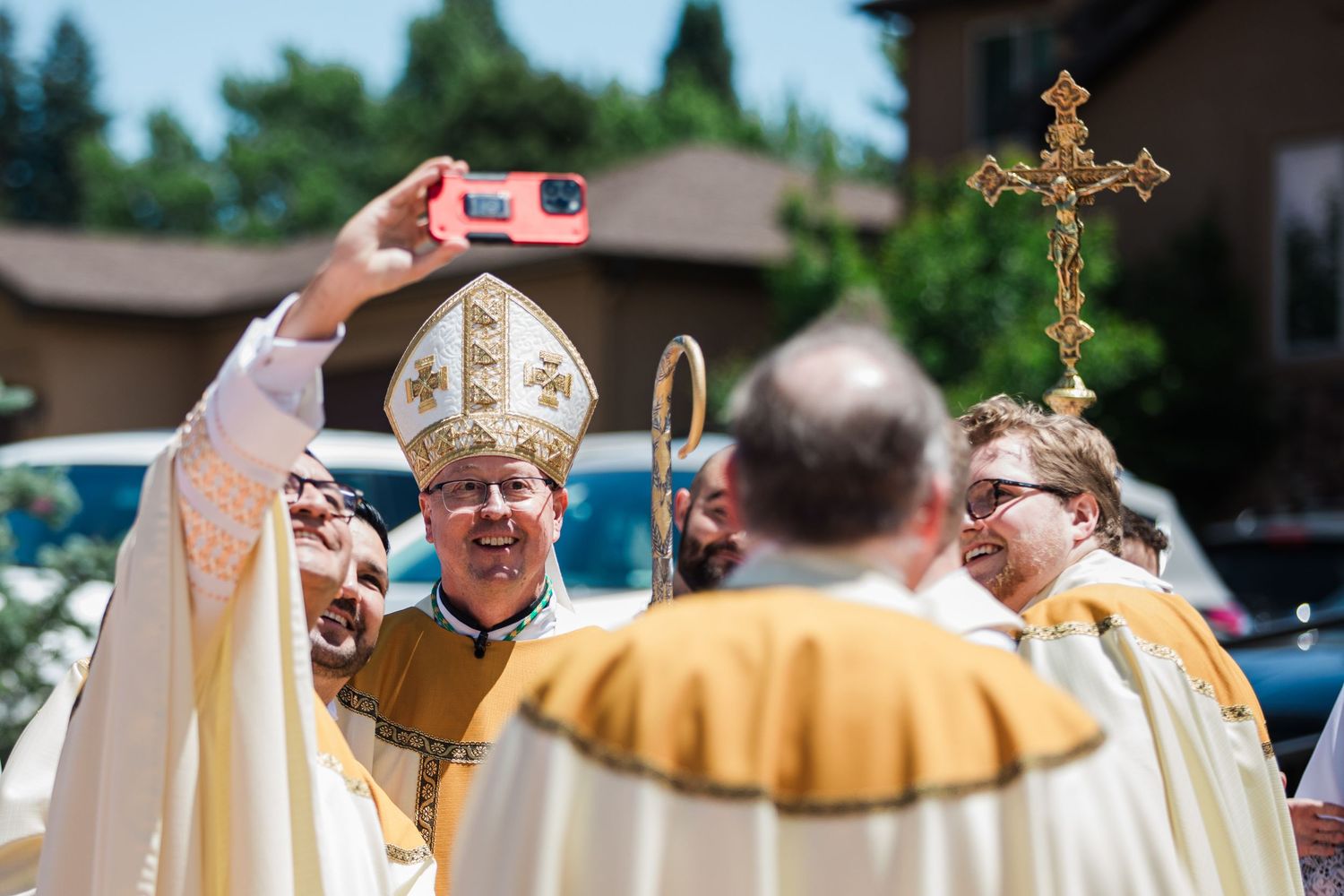 Catholic bishops in ceremonial robes take a selfie outdoors during a religious procession with a golden cross.