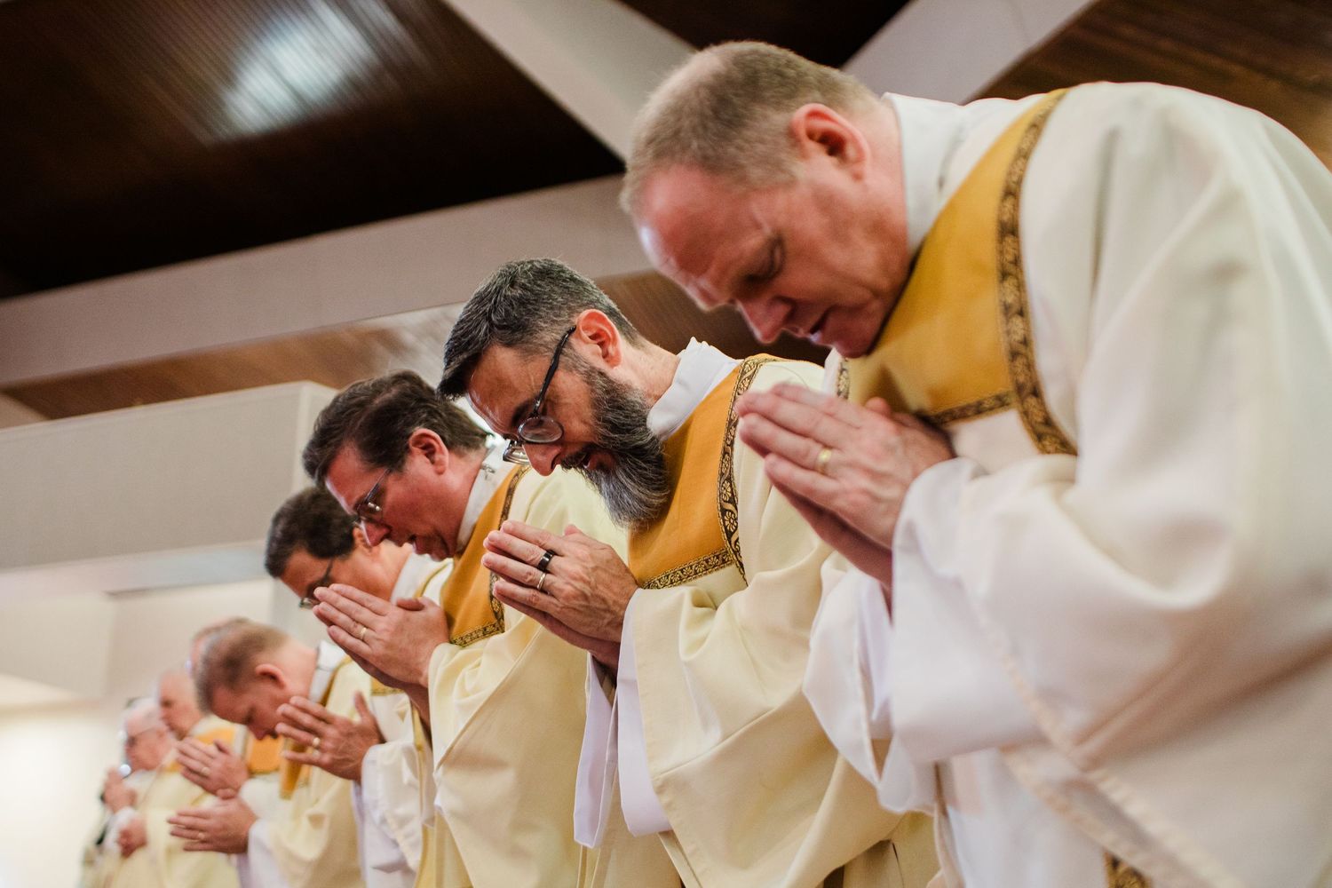 Catholic priests in white vestments bow their heads in prayer during an ordination ceremony.
