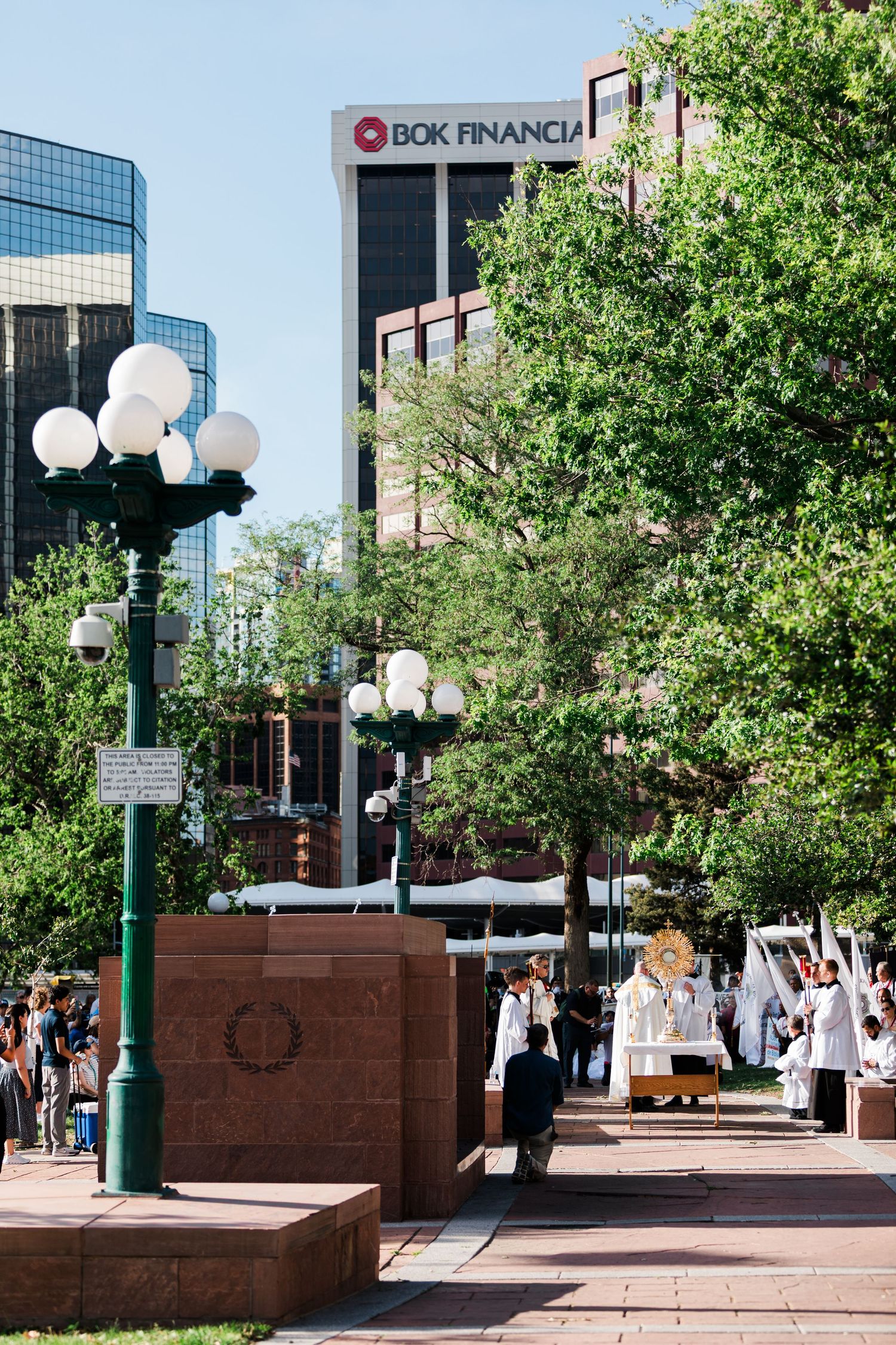 People gather in a plaza near BOK Financial building, surrounded by green trees and decorative lamp posts on a sunny day.