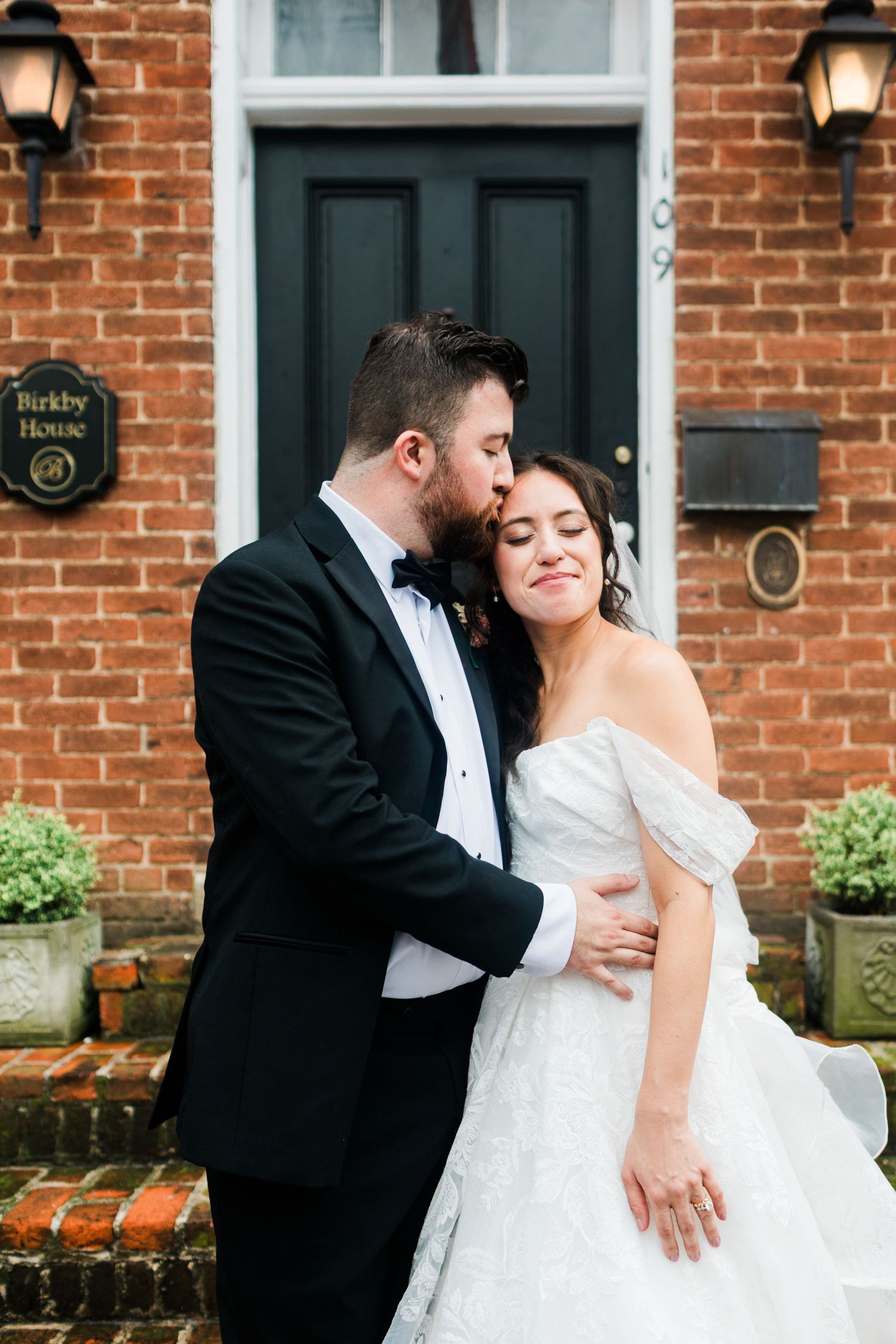 Groom in black tuxedo kisses bride in off-shoulder wedding gown outside Birkby House brick building.