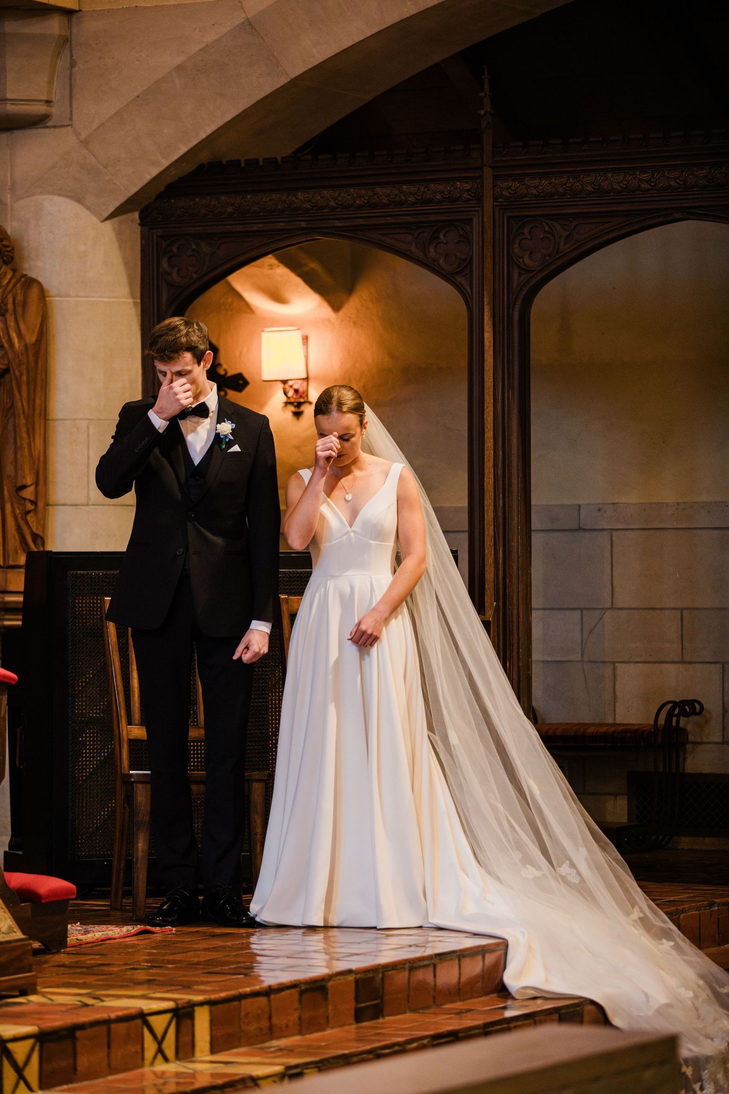 Emotional bride and groom wiping tears during wedding ceremony in elegant chapel with warm lighting.