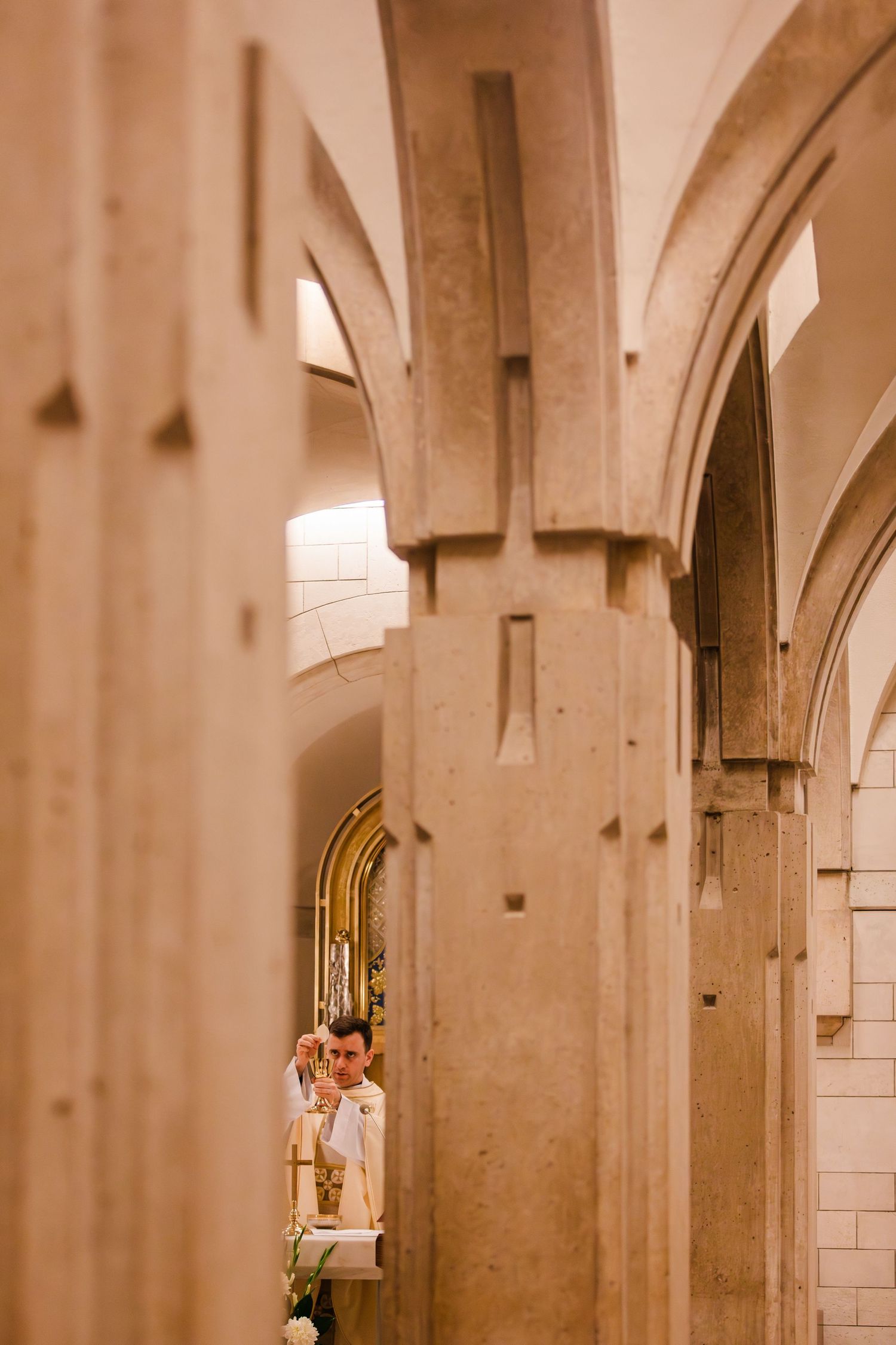 Priest performing a ceremony at a church altar, viewed through ornate stone arches of a cathedral.