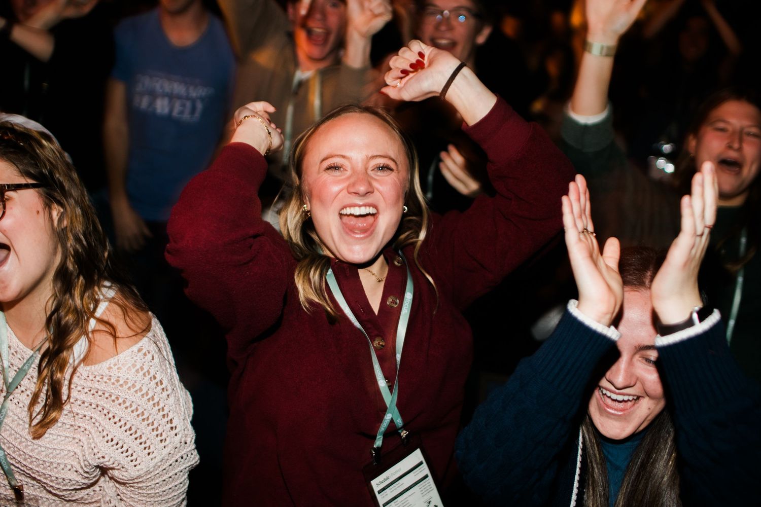 A crowd of excited people at a concert or event cheering with their hands raised in the air.
