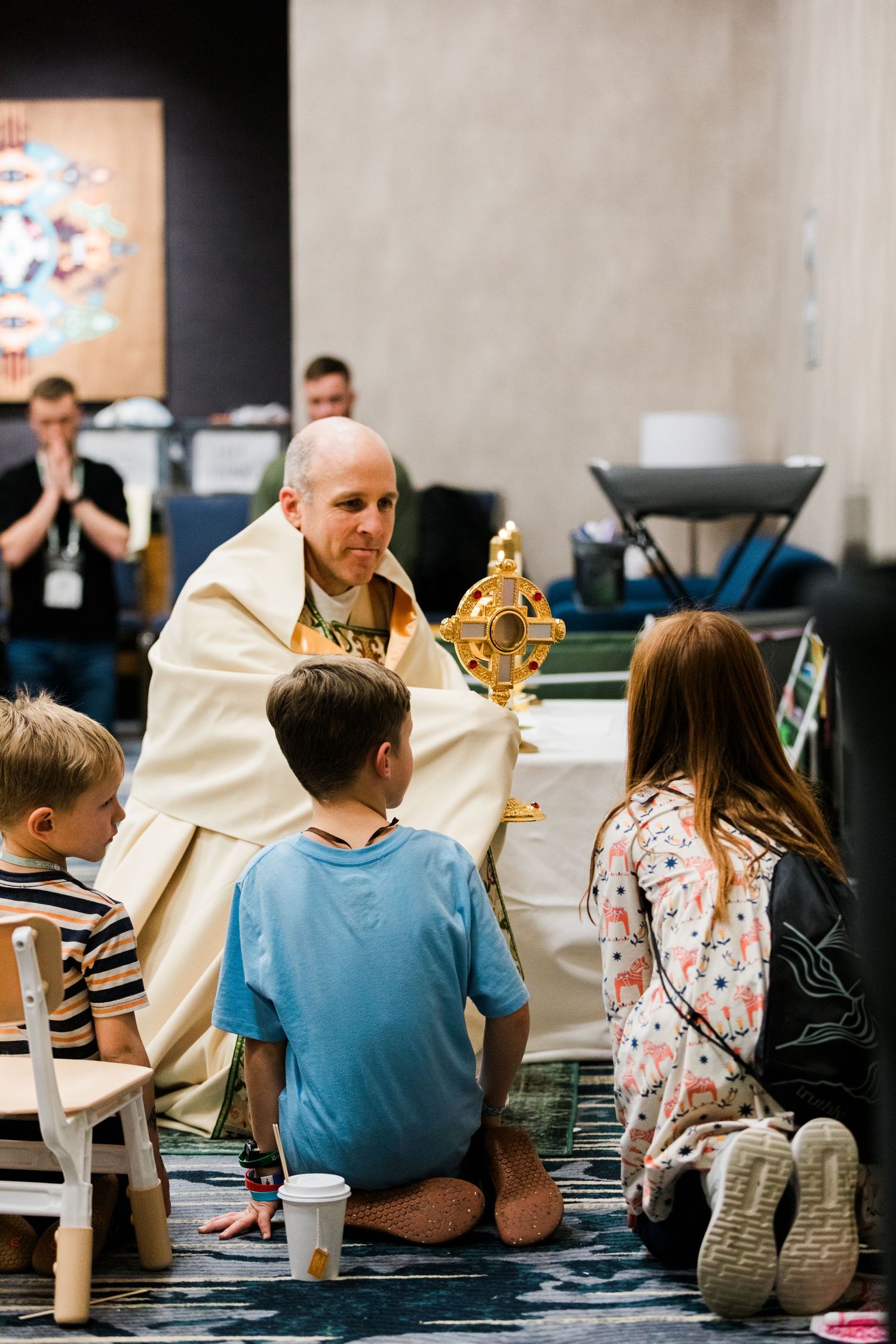 A priest in white robes holds a cross while speaking to a group of children seated in a church setting.