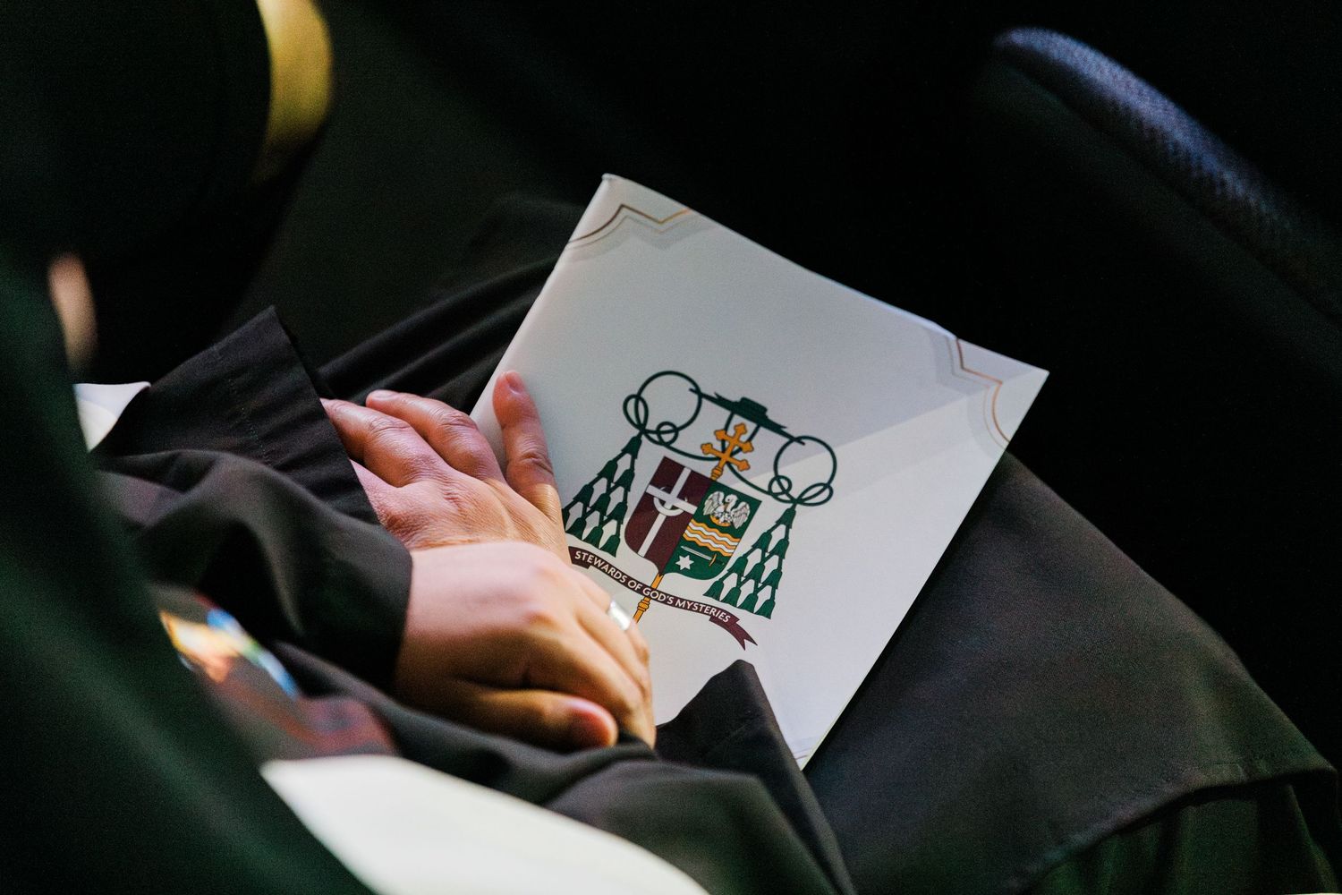 Attendee holding Catholic bishop ordination program booklet featuring episcopal coat of arms at ceremony.