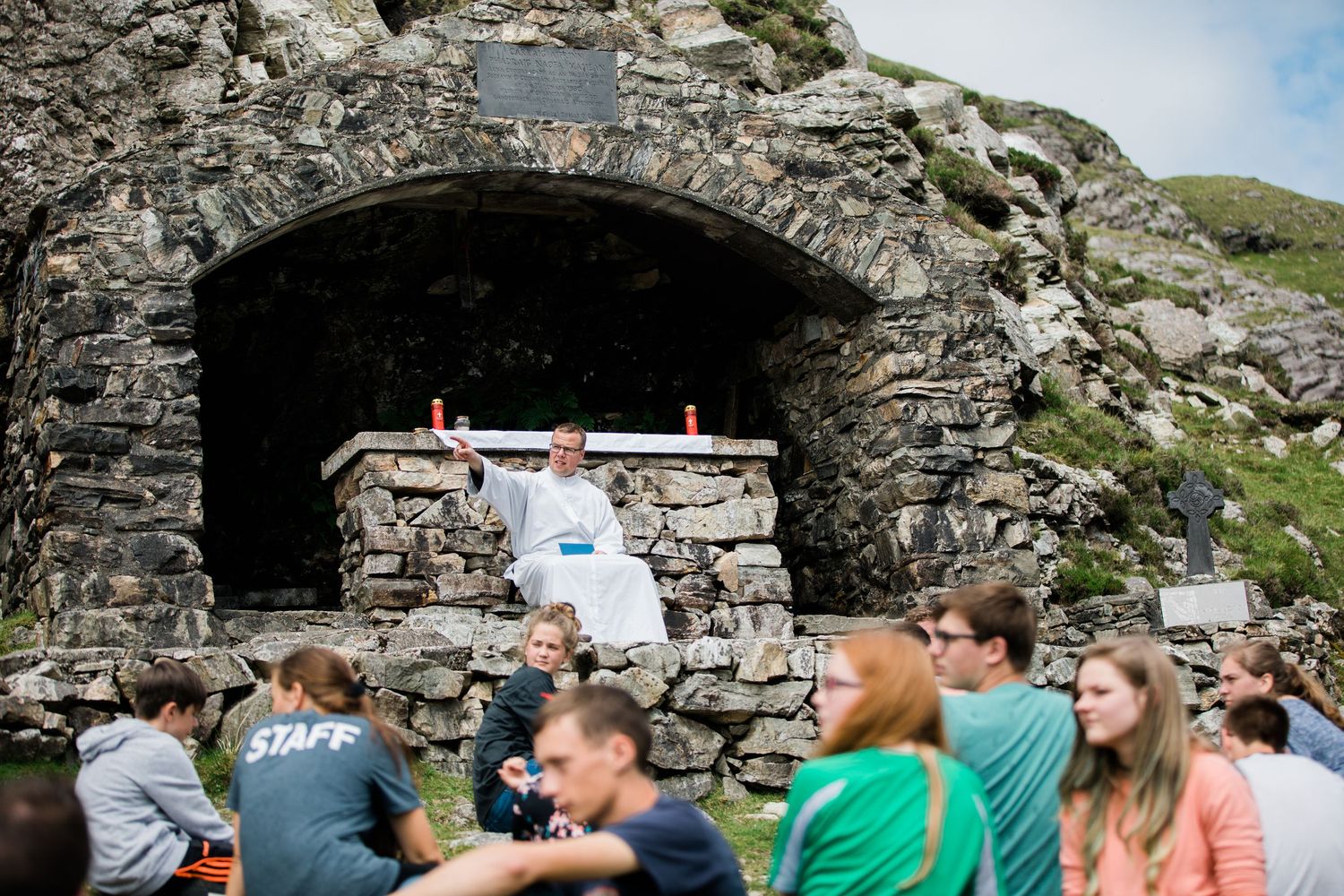 Priest celebrating outdoor Mass at a stone grotto altar surrounded by young pilgrims in Ireland.