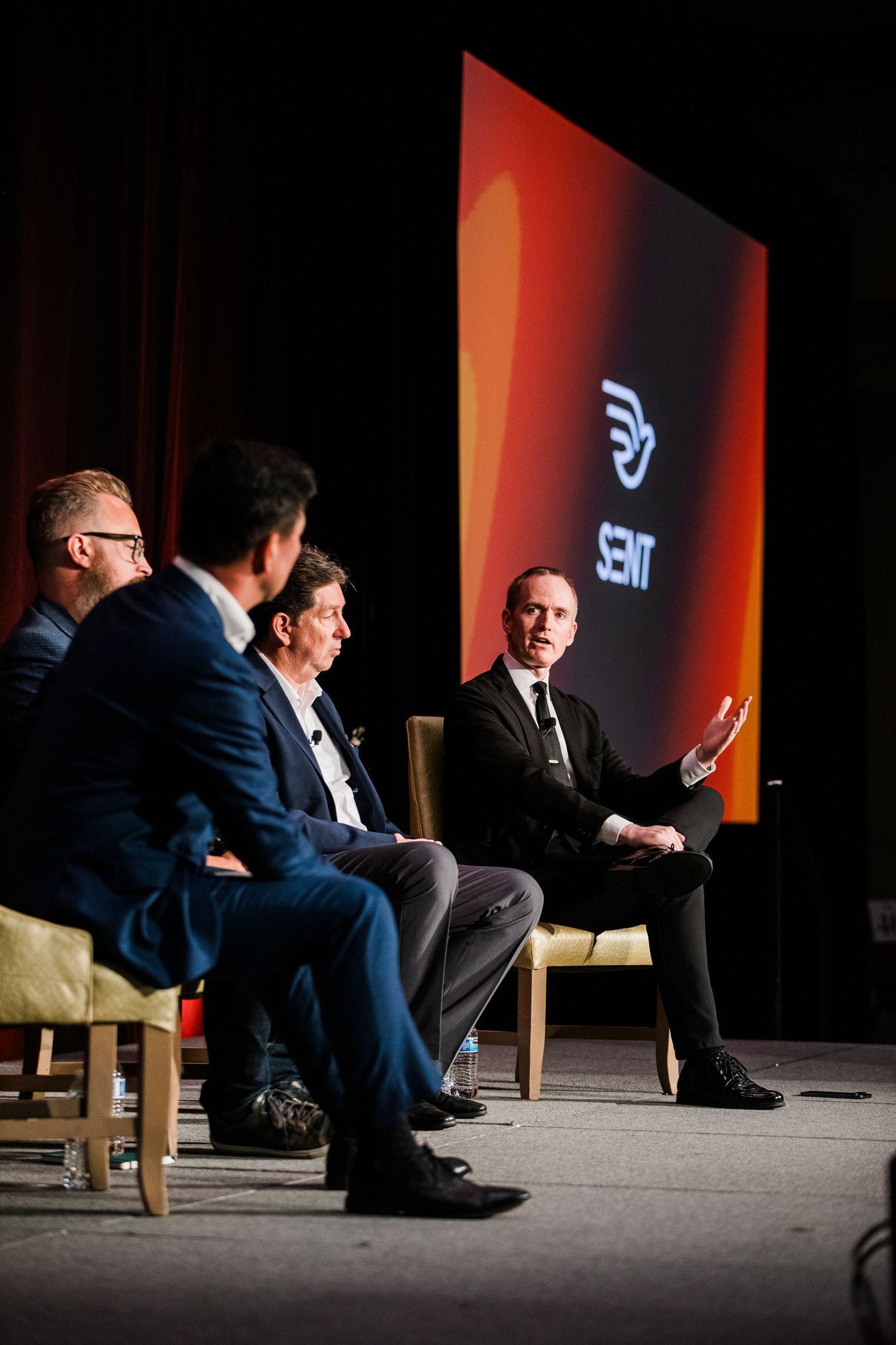 Four professionals in suits participate in a panel discussion on stage with a SENT logo projected on screen behind them.