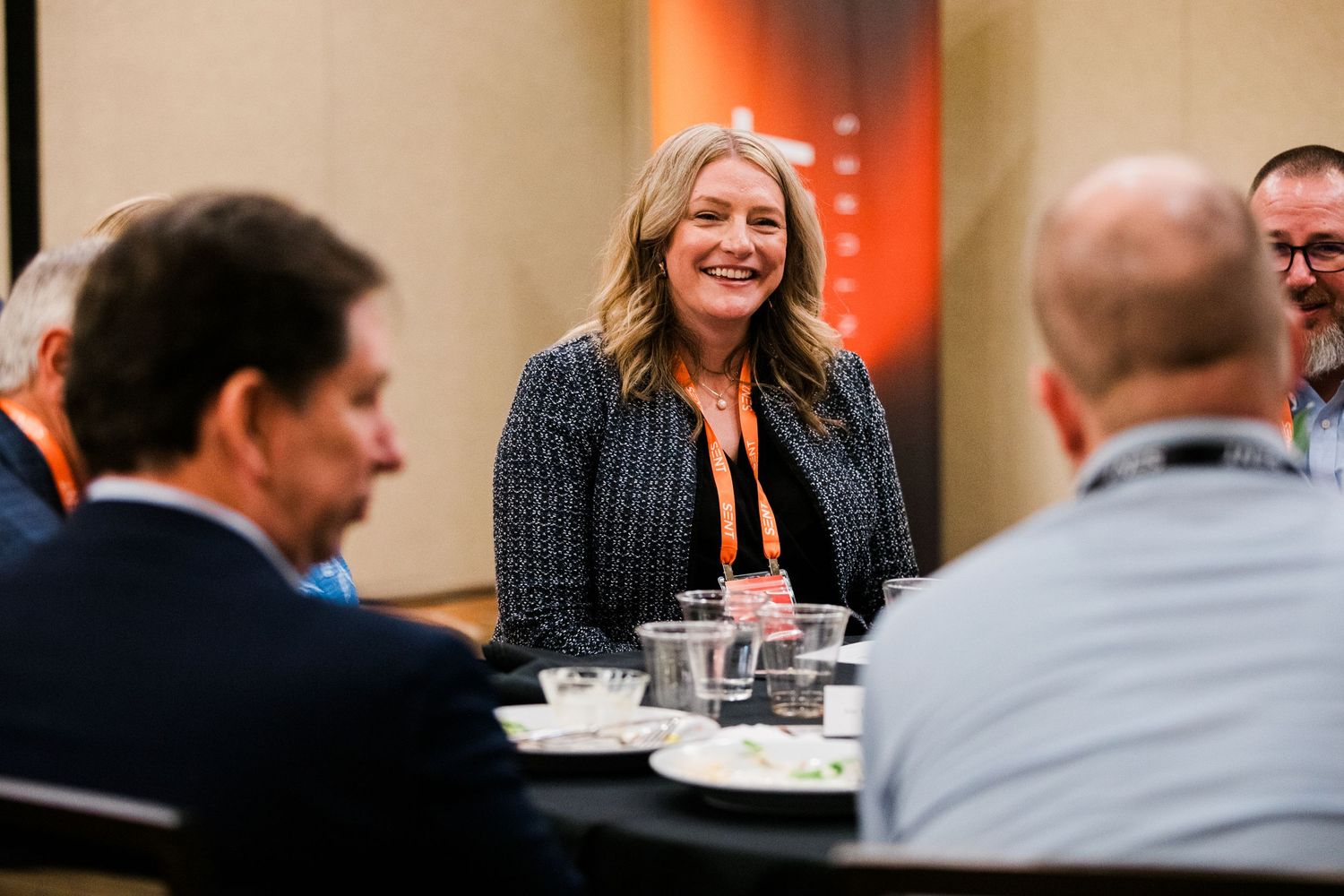 Smiling woman in blazer at conference networking lunch table with colleagues.
