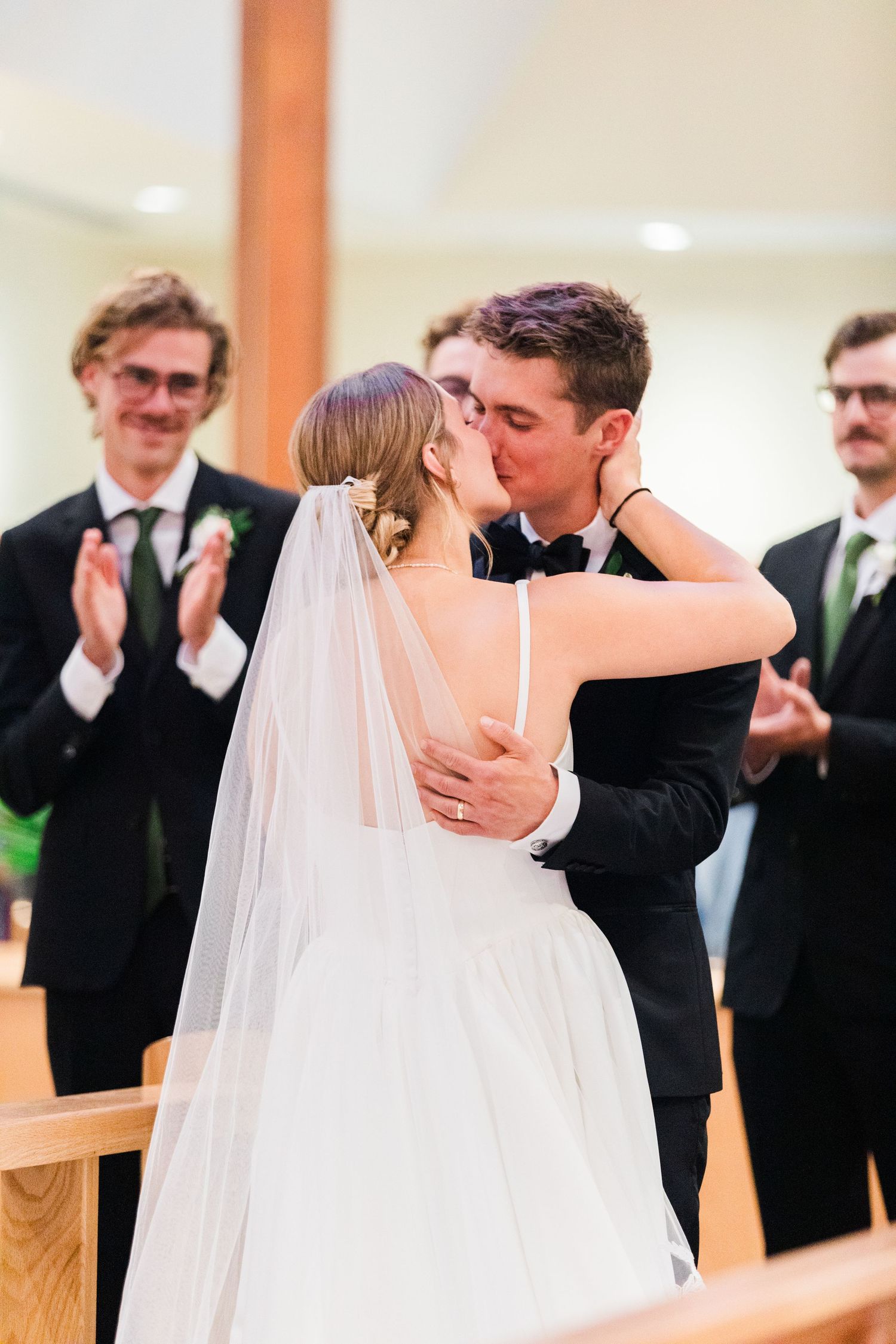 Wedding couple shares intimate first kiss during ceremony while guests applaud in background.