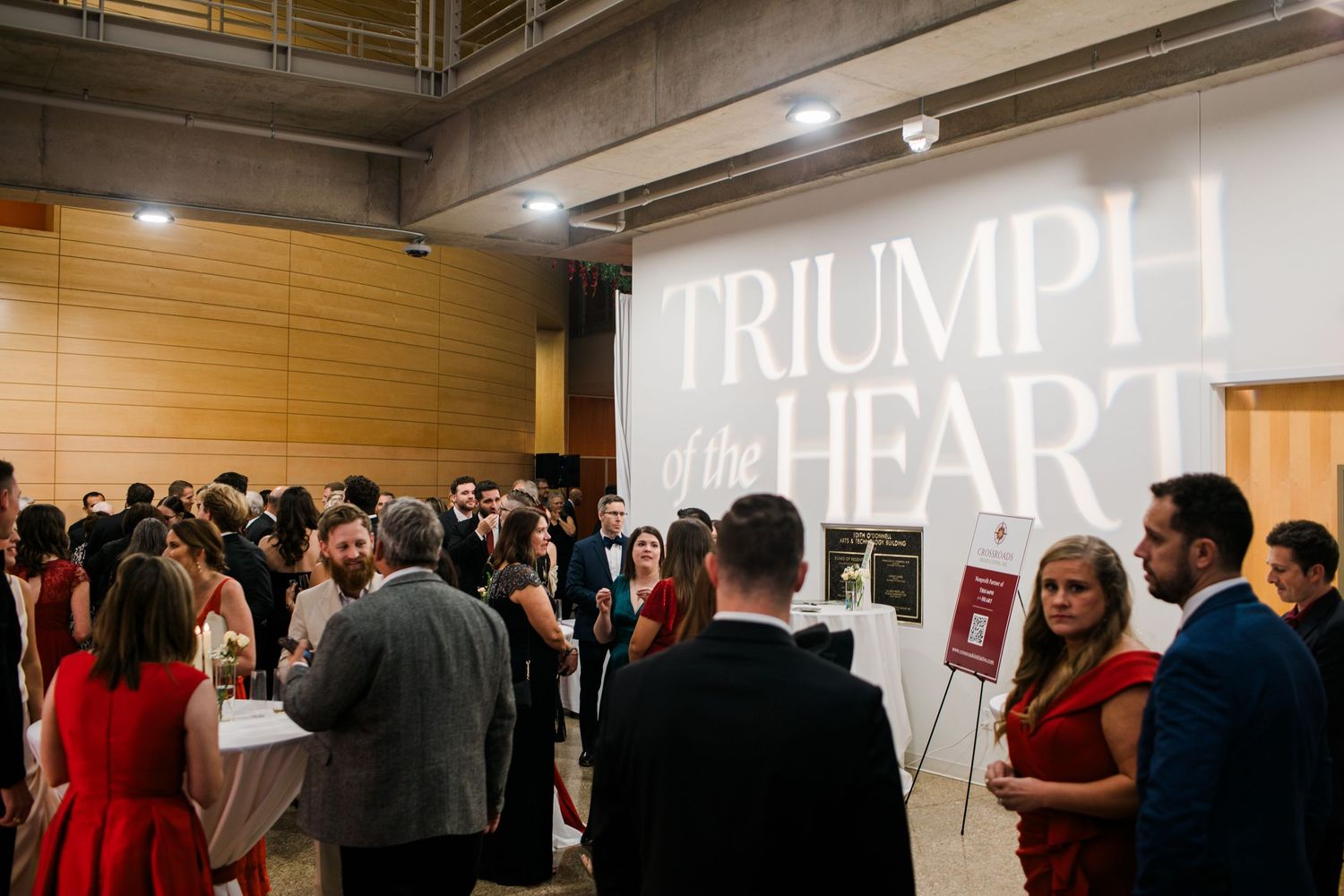 Crowd gathered at an art exhibition with large text reading 'TRIUMPH OF HEART' on the wall.
