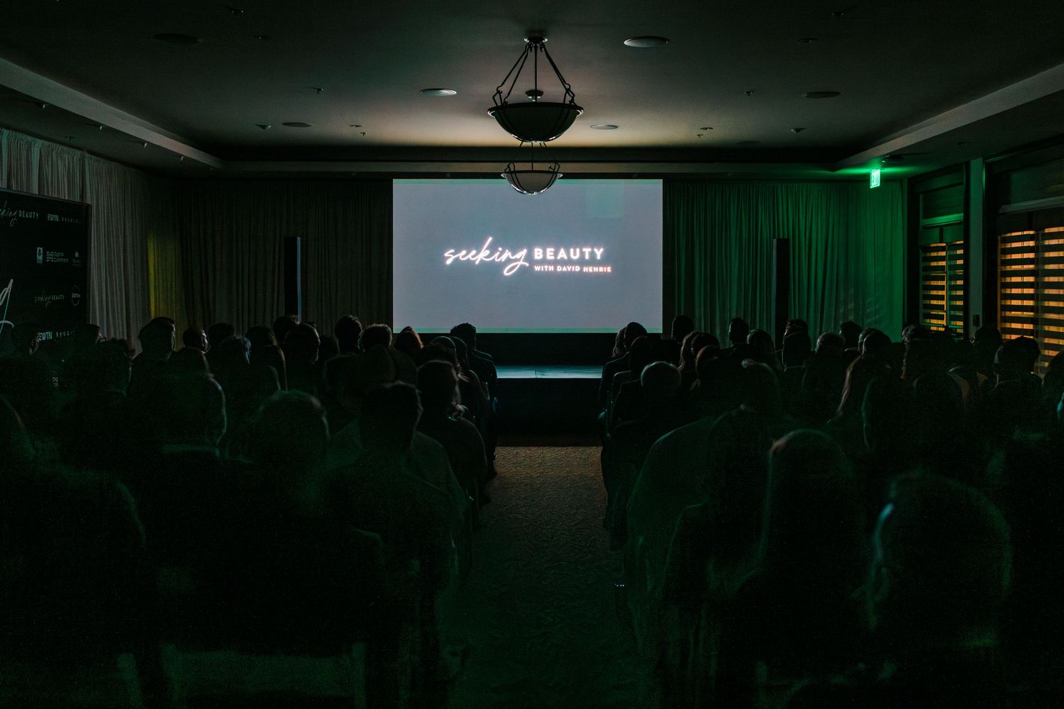 A dark auditorium filled with silhouettes of people watching a presentation with green ambient lighting.