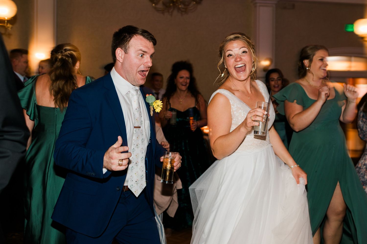 Bride and groom dancing joyfully at wedding reception, guests in emerald green dresses celebrating in background.