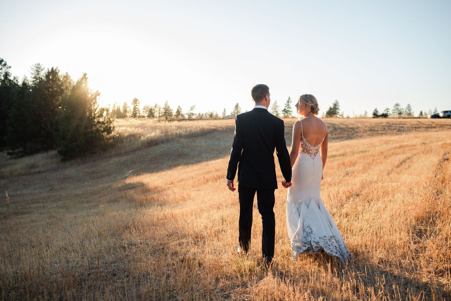Bride and groom holding hands walking through golden field at sunset, bride in backless lace gown.