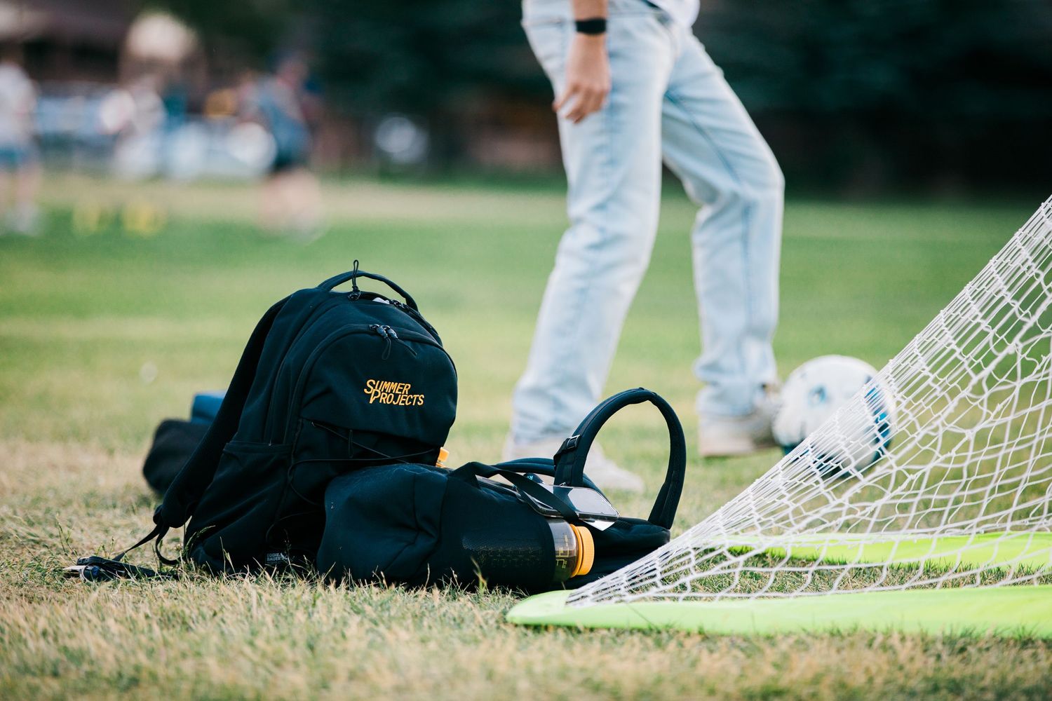 Black backpack and soccer goal on grass field with player and ball in background.