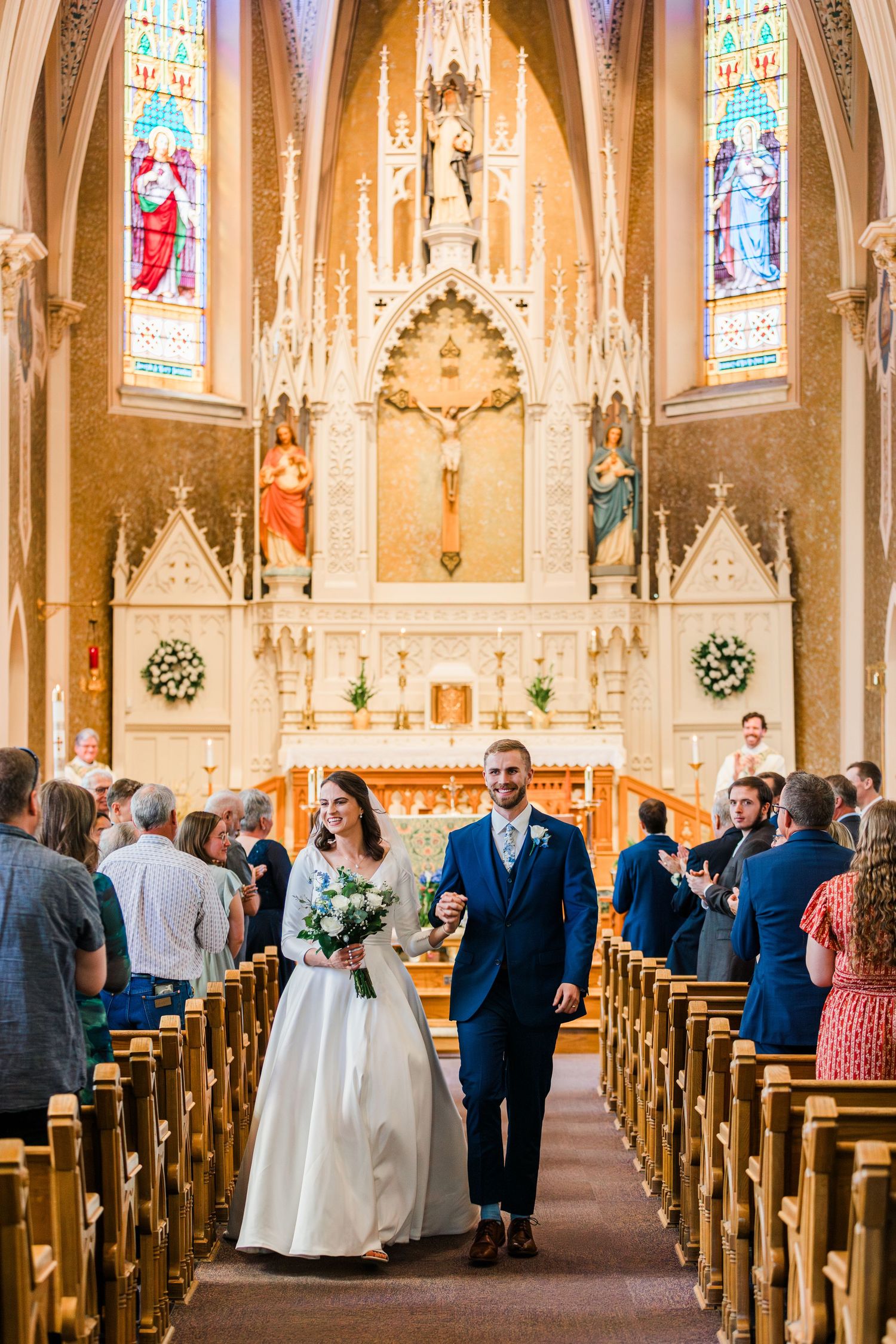 Bride and groom walk down church aisle after wedding ceremony, with ornate altar and stained glass windows behind them.