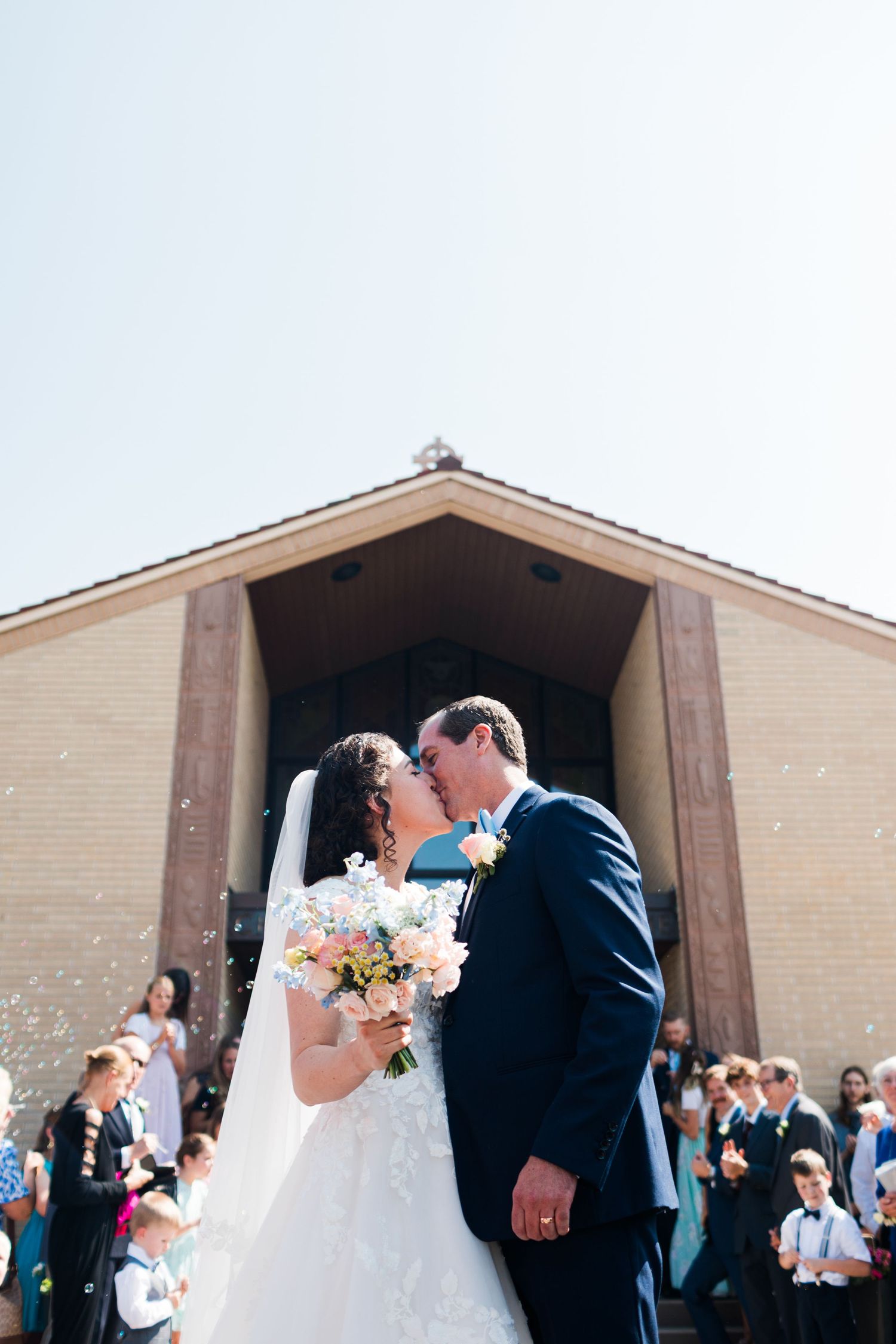 Bride and groom share first kiss outside church after wedding ceremony, bride holding pastel bouquet.