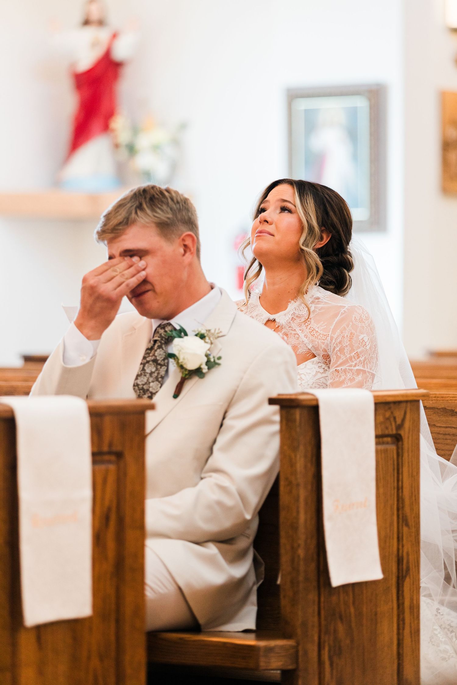 Emotional groom wipes tears while bride looks up during their Catholic church wedding ceremony.
