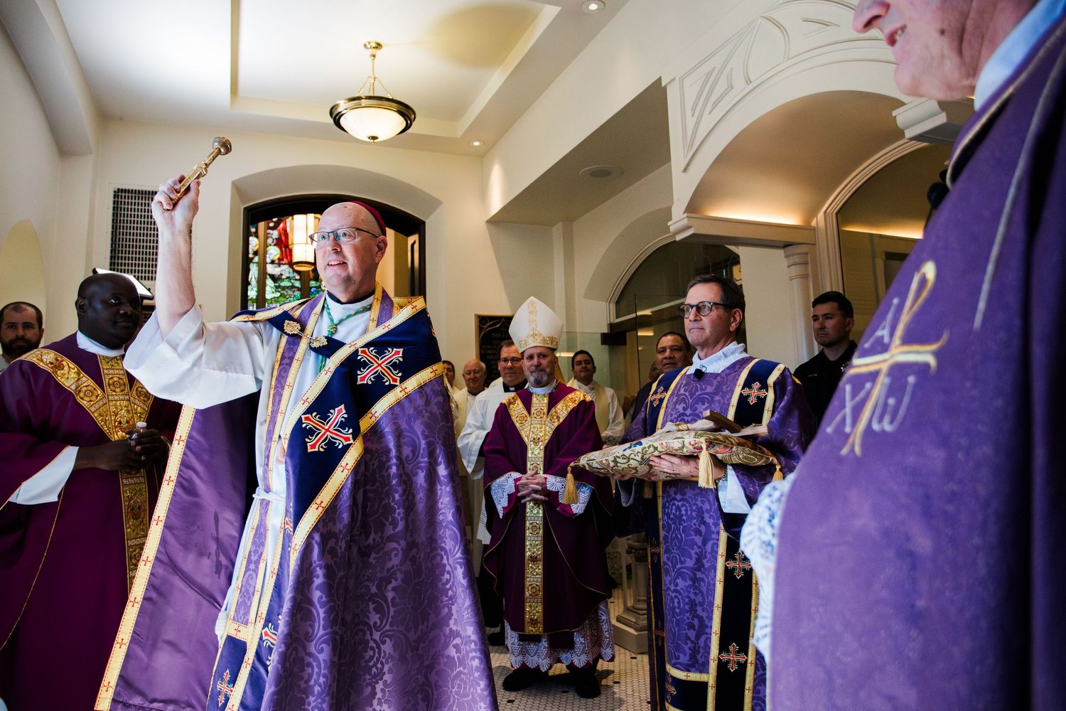 Clergy members in purple vestments with gold crosses perform a religious ceremony in an ornate church hallway.