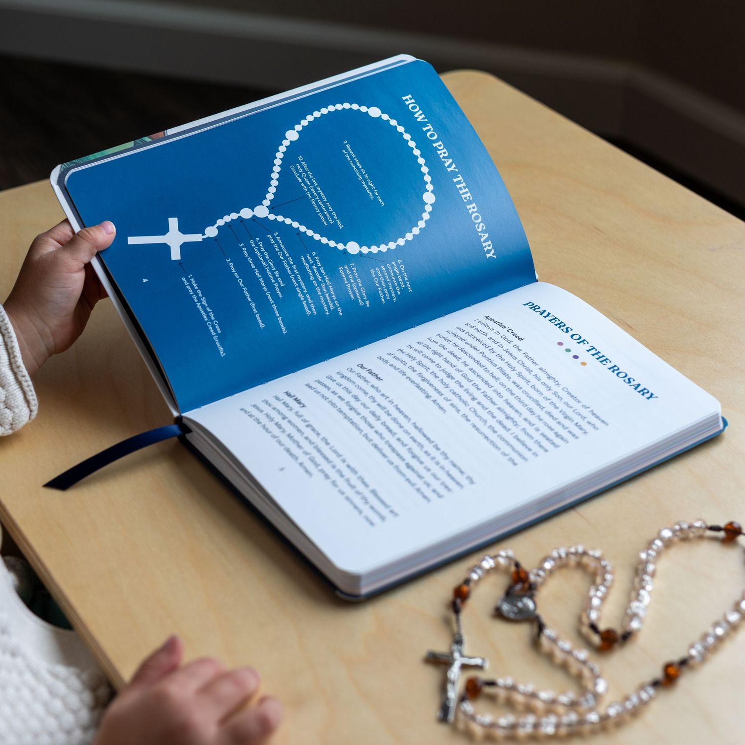 Open rosary prayer book on a wooden desk with a Catholic rosary beaded necklace beside it.