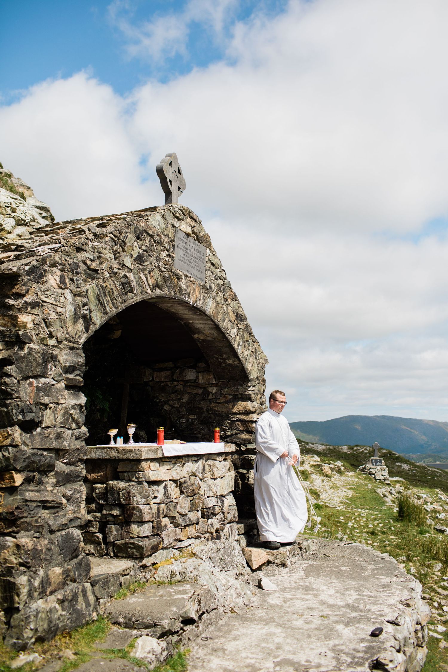 Priest in white robes stands beside a stone altar with Celtic cross atop a mountain shrine under cloudy skies.